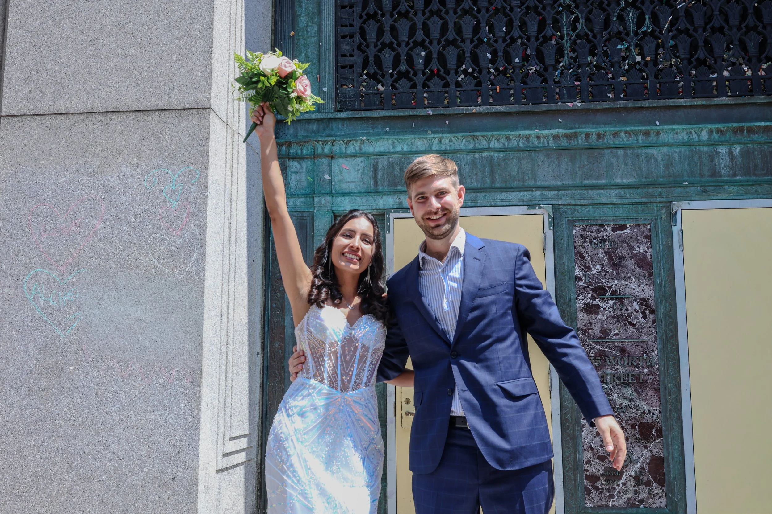 A smiling man and woman in wedding attire posing outdoors. The woman, wearing a sparkle wedding dress, holds a bouquet of pink and white roses above her head. The man, in a blue suit, stands beside her with his arm around her waist, both looking happ