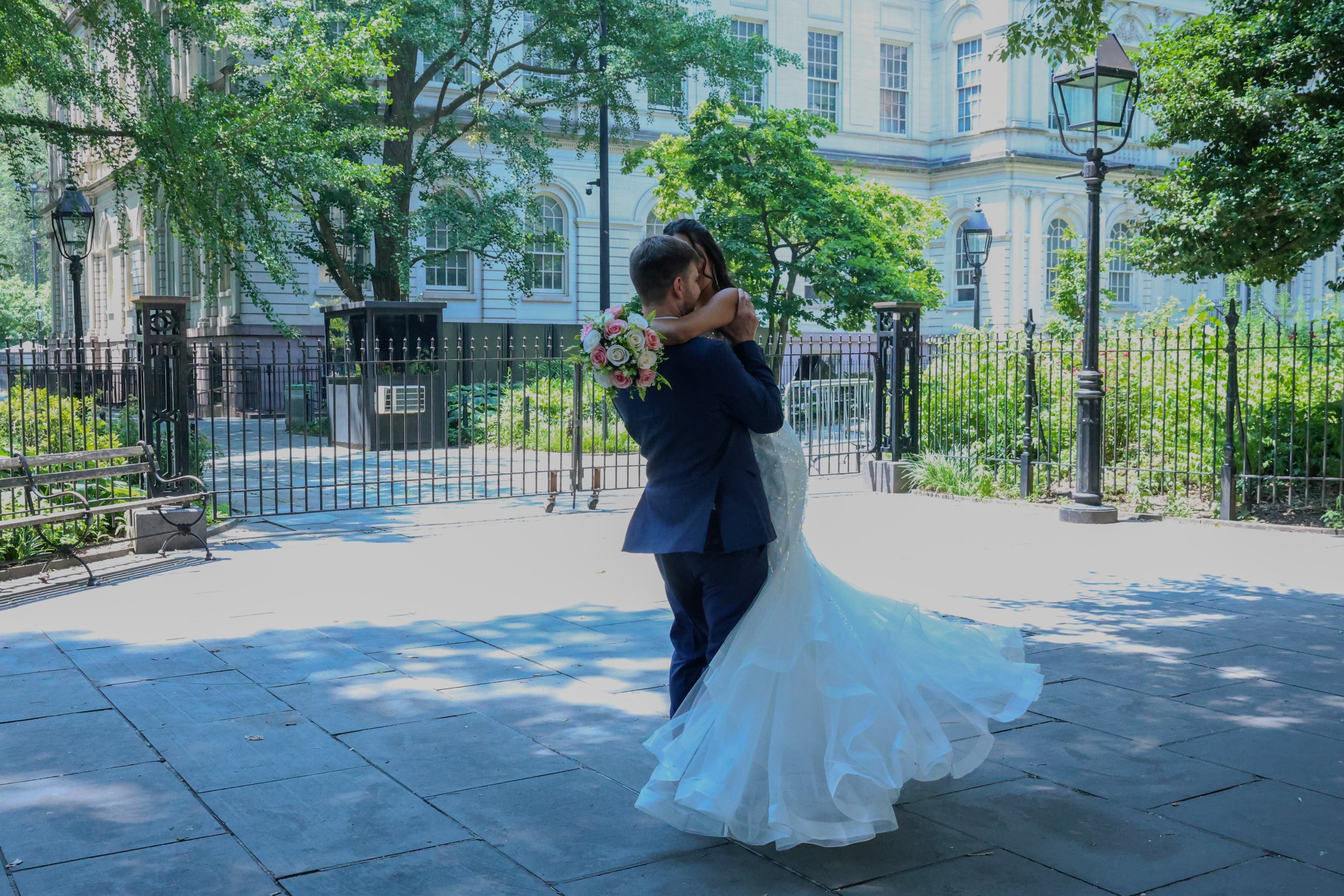A newlywed couple embracing and kissing outdoors in front of a historic building, with the bride holding a bouquet of pink and white roses. The groom is wearing a dark suit, and the bride is in a white wedding gown.