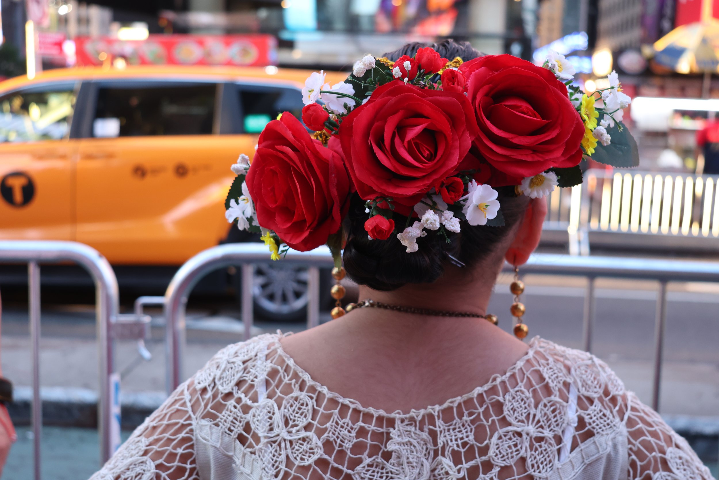 A woman with dark hair styled in a bun decorated with a large red floral headpiece, wearing gold earrings and a white, open-weave crochet top, standing in front of a yellow taxi cab on a busy city street at dusk.