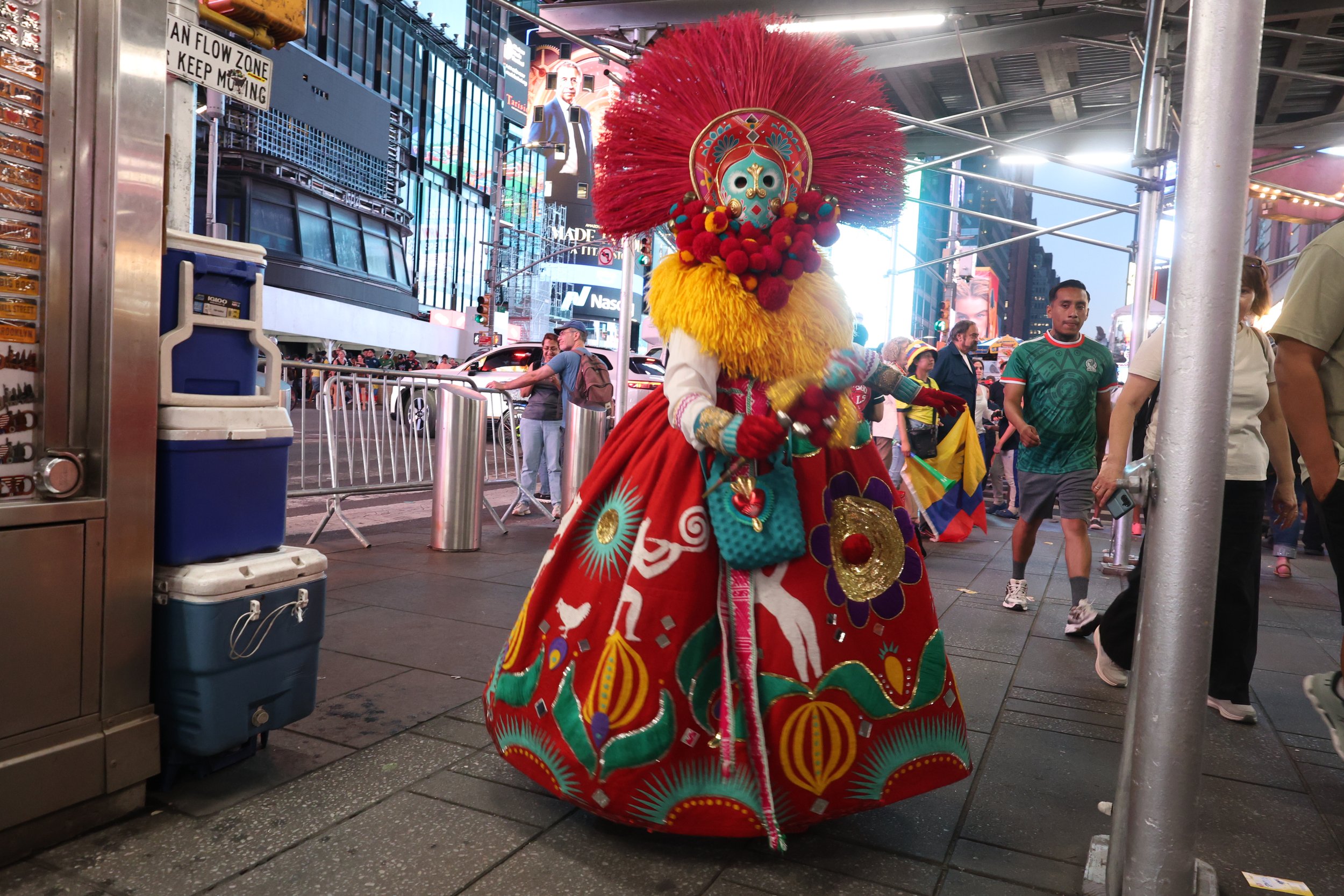 A performer dressed in a colorful traditional costume with a decorated mask and large red headdress, walking in a busy urban area at night with bright billboards and people in the background.