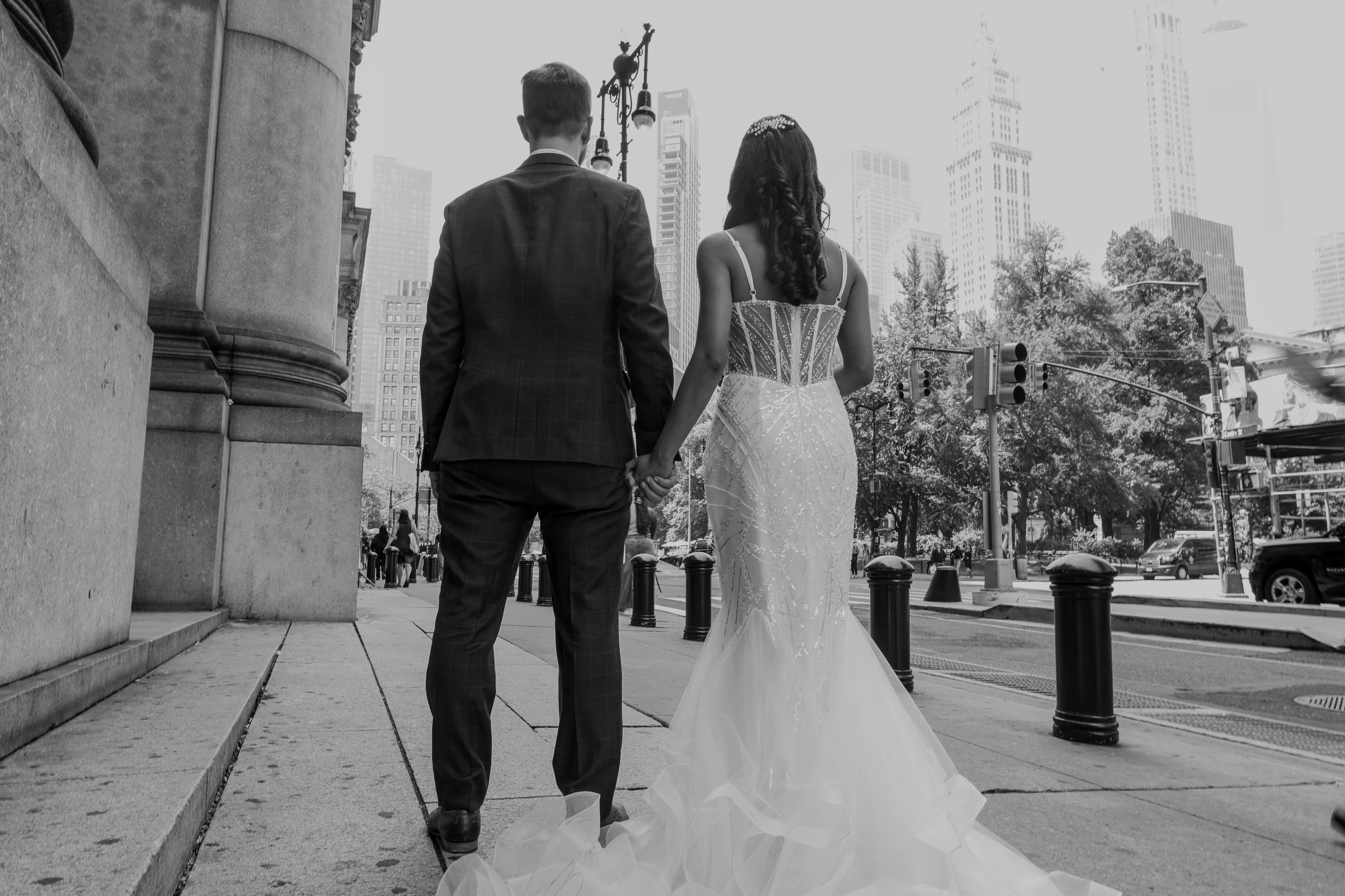 A man and woman holding hands walking away in an urban setting, with tall buildings and trees in the background, black-and-white photo.
