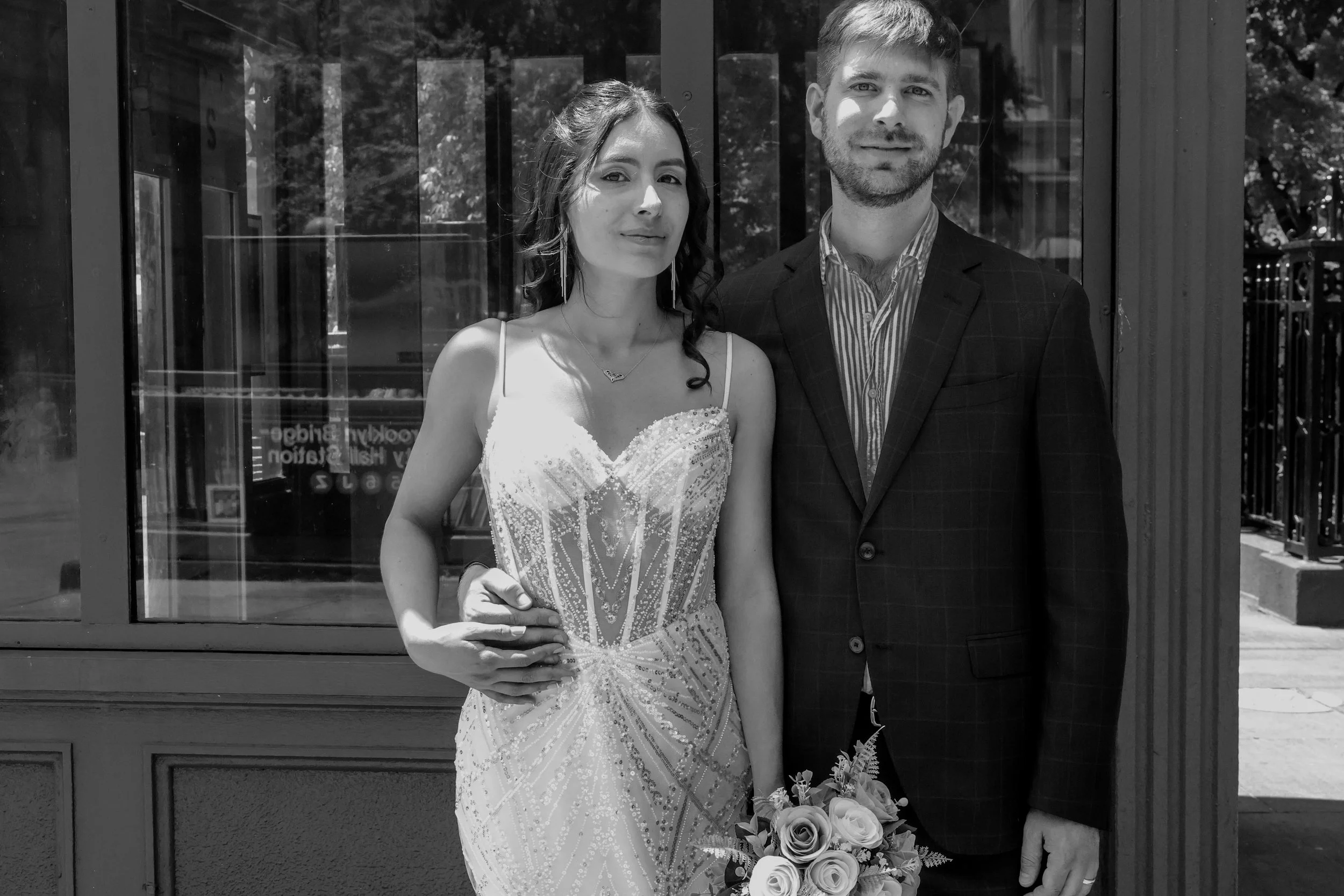 A black and white photo of a couple, the woman in a sleeveless, embellished gown holding a bouquet of roses and the man in a suit, standing outside in front of a glass window.