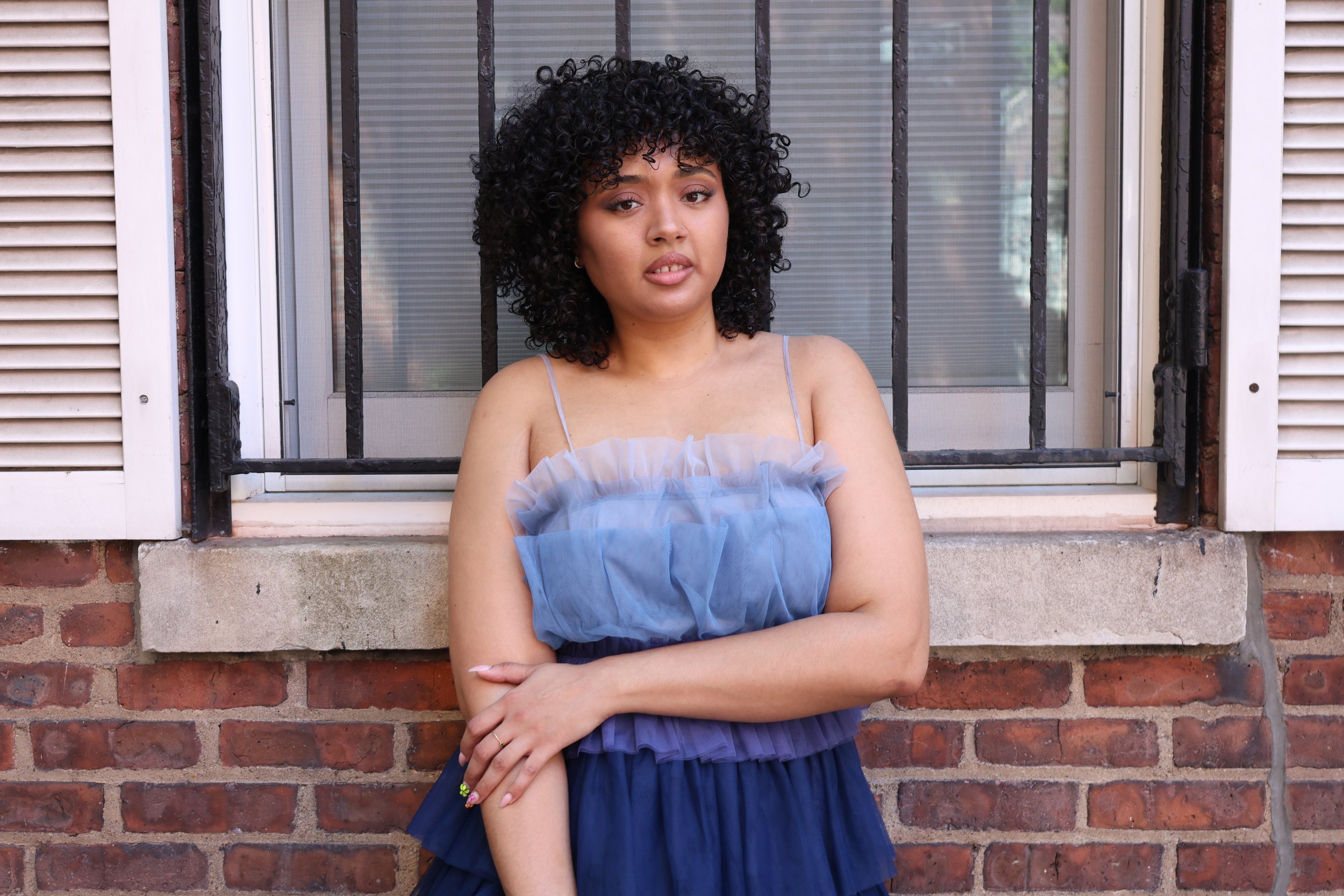 A woman with curly black hair standing in front of a brick wall and a window with white shutters and black metal bars, wearing a blue strap dress with ruffles, arms crossed, looking at the camera.