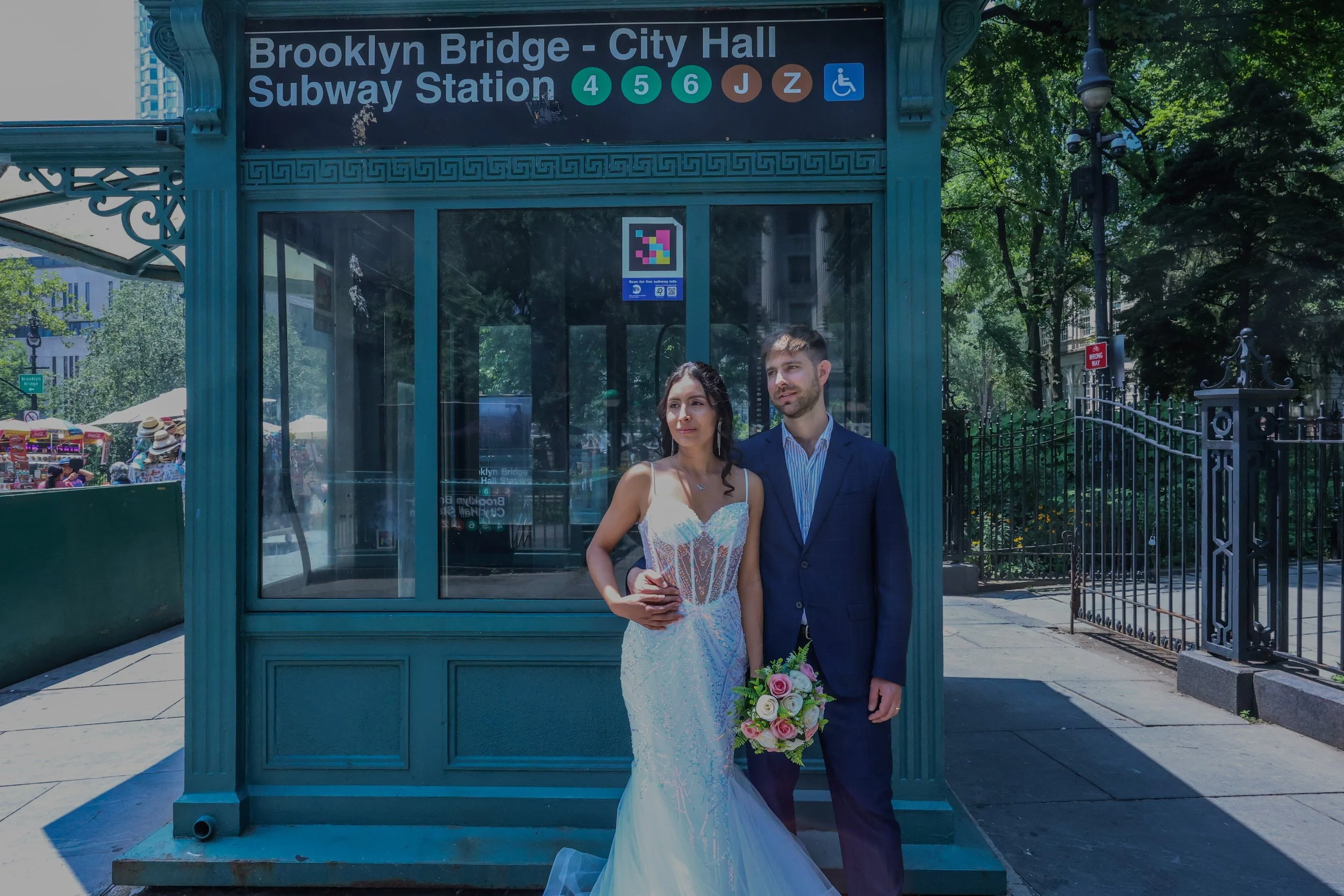 A couple dressed in wedding attire standing together in front of a subway station entrance. The woman is holding a bouquet of flowers. The station sign indicates Brooklyn Bridge - City Hall Subway Station, with icons for multiple subway lines and acc