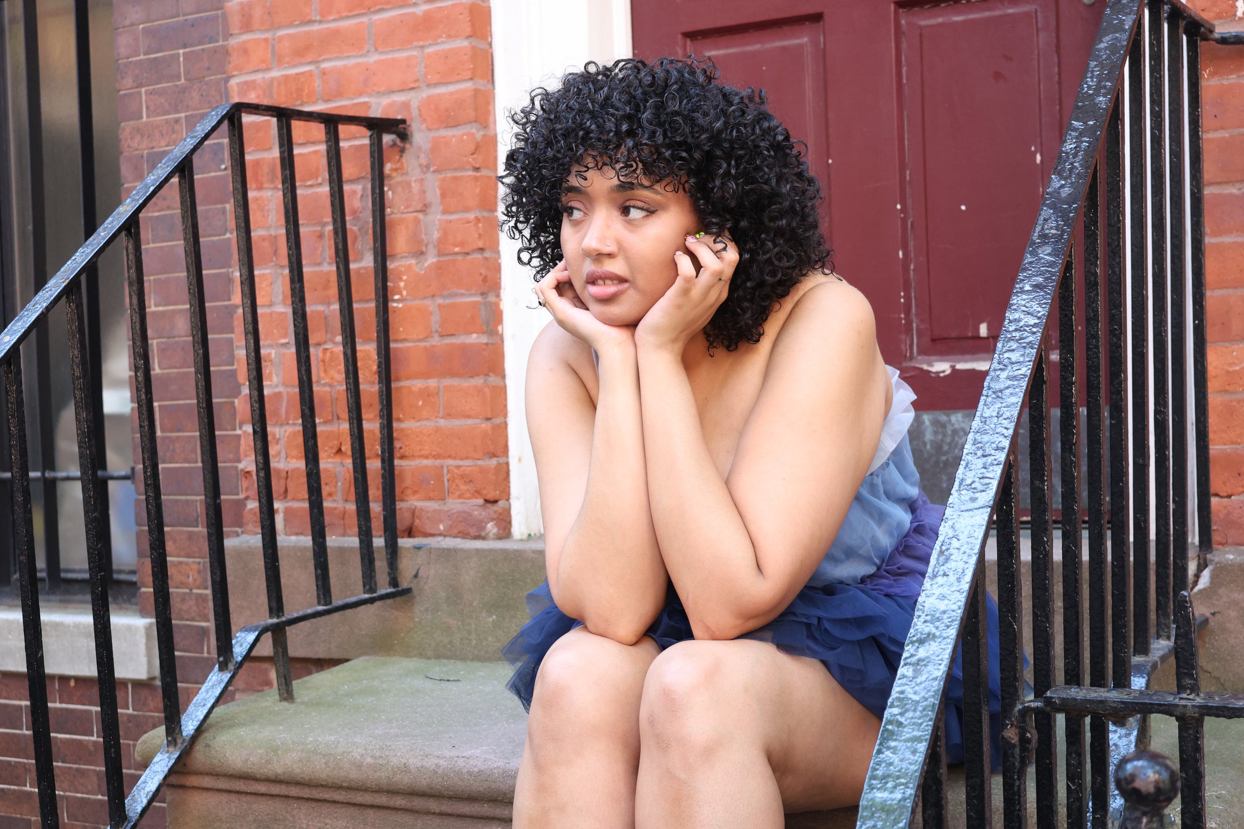 A young woman with curly black hair sitting on a concrete step outside a brick building, resting her face in her hands, looking to the side, wearing a strapless top and a blue skirt.