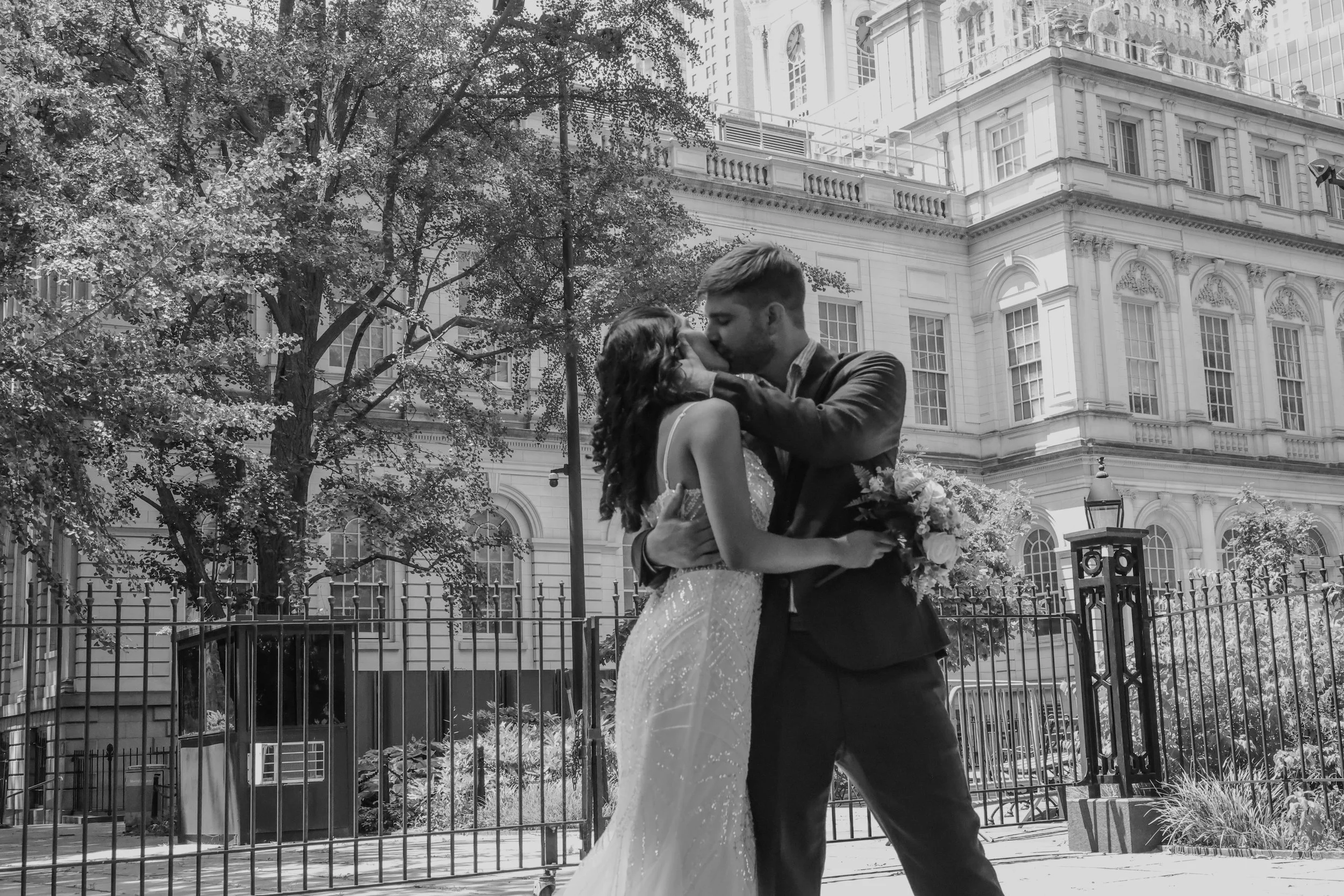 A black and white photo of a couple sharing a kiss, with the woman wearing a wedding dress and holding a bouquet, and the man dressed in a suit, set against a backdrop of a historic building and trees.