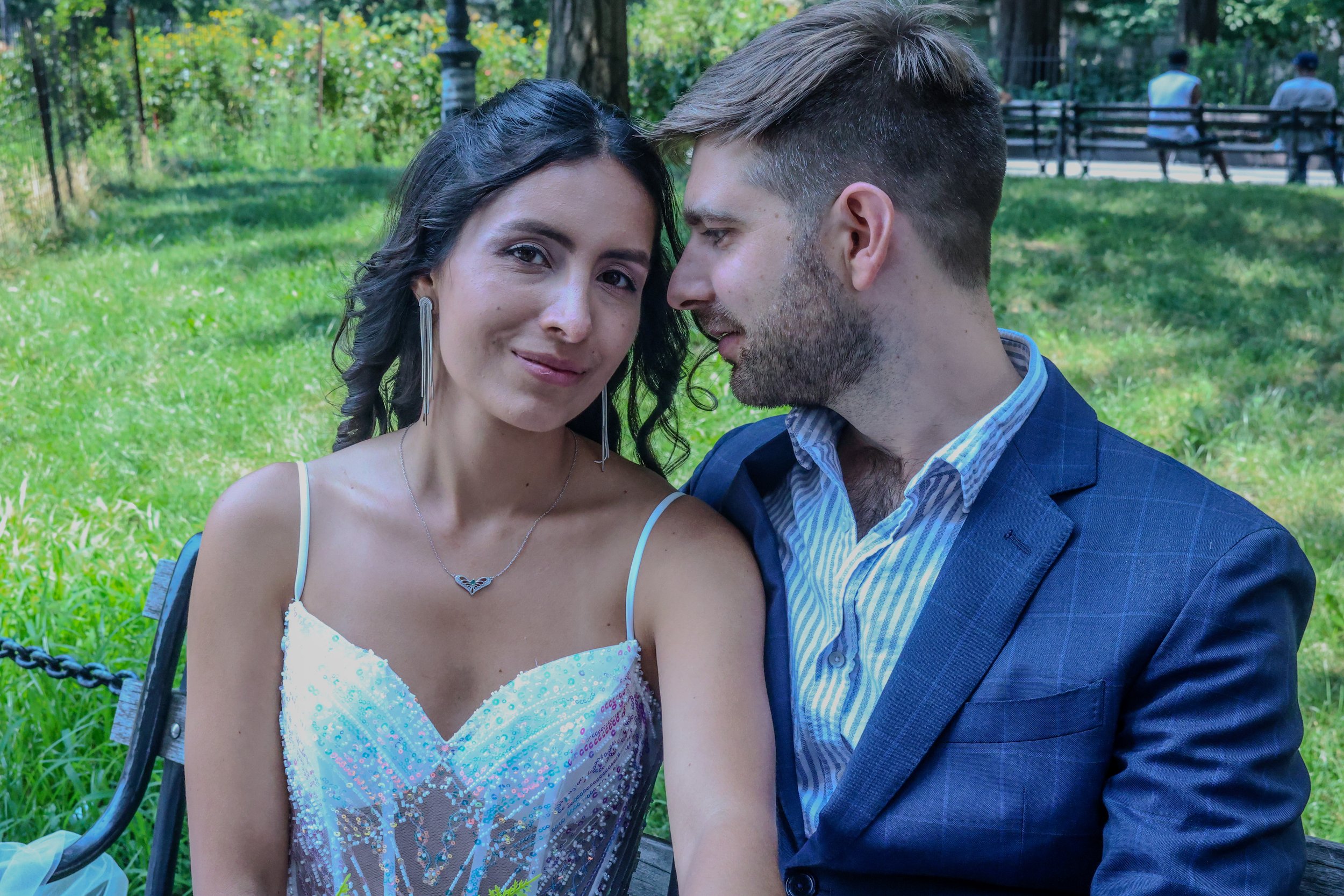 A young woman and a young man sitting closely together outdoors, with the woman smiling slightly at the camera and the man looking at her, in a park with green grass and trees in the background.