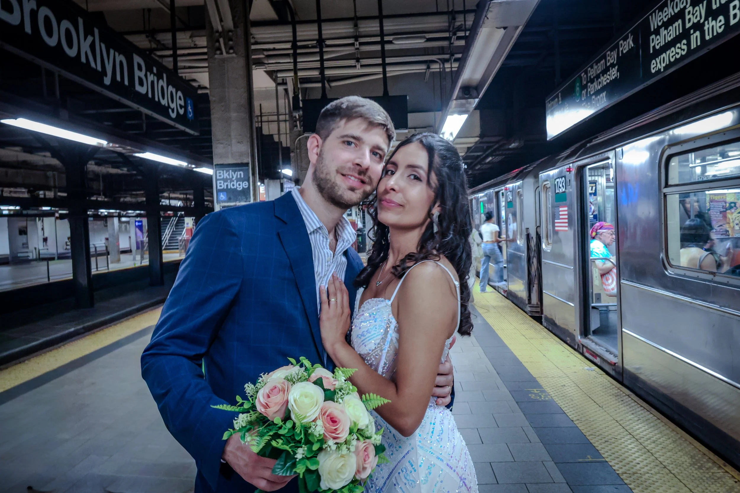 A newlywed couple, dressed in wedding attire, stands close together on a subway platform at Brooklyn Bridge station. The groom is wearing a blue suit and the bride is in a white wedding dress with intricate blue details. The bride holds a bouquet of 