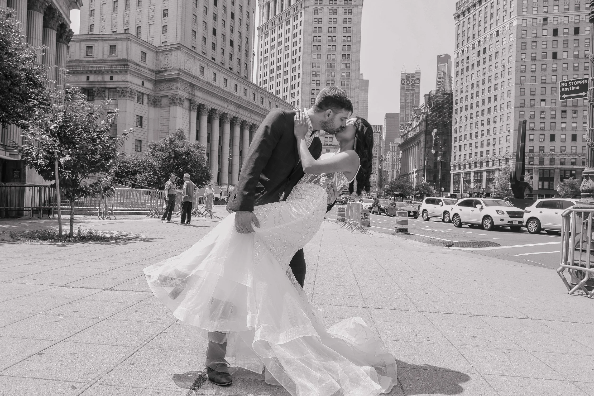 A couple in wedding attire sharing a kiss in an urban city square with tall buildings in the background.