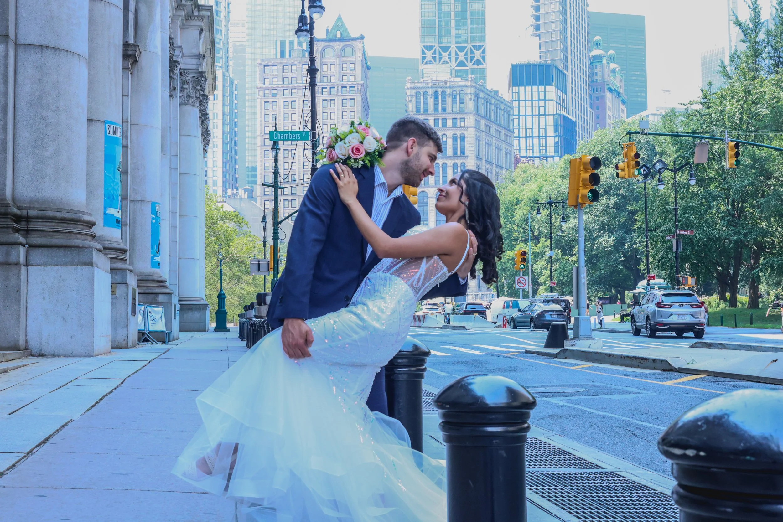 A couple dressed in wedding attire, with the woman in a white wedding gown holding a bouquet of flowers, sharing an intimate moment on a city street with tall buildings, traffic lights, and parked cars in the background.