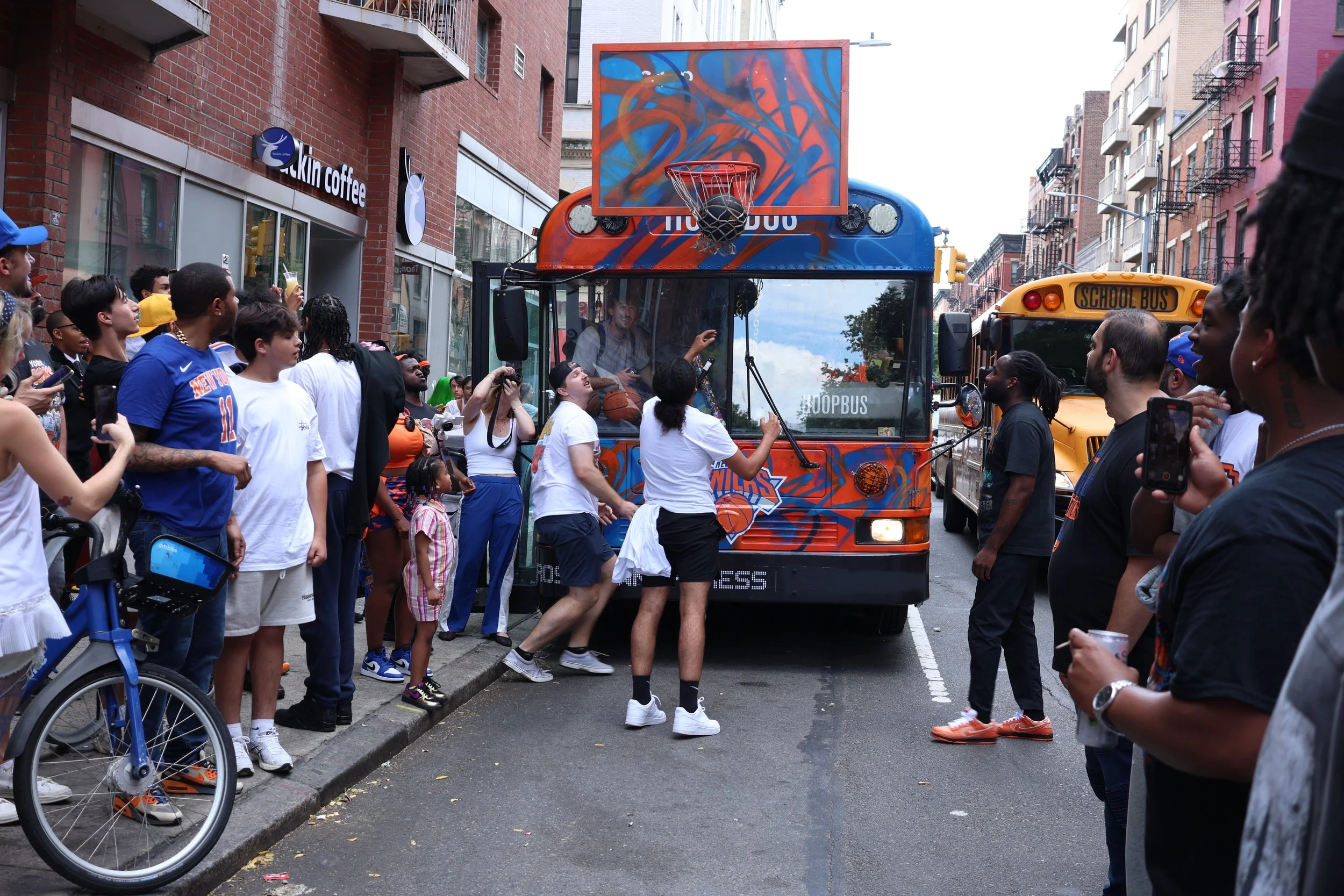 Crowd of people gathered around a small basketball-themed streetcar on a city street, with buildings and a school bus in the background.