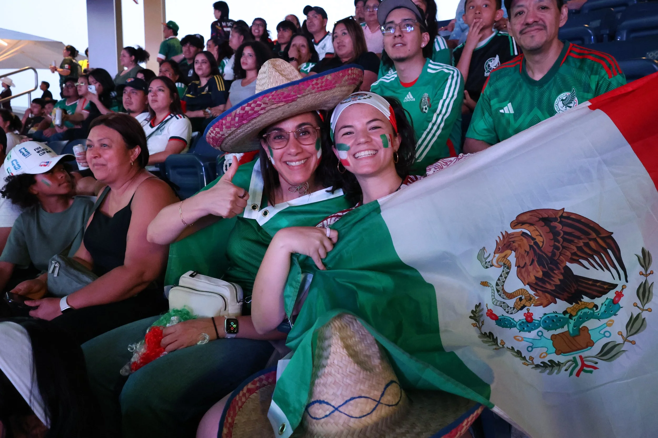 A group of diverse sports fans in the stands, wearing Mexican team jerseys and accessories, with two women in the foreground smiling and holding a Mexican flag. They are at a sporting event.