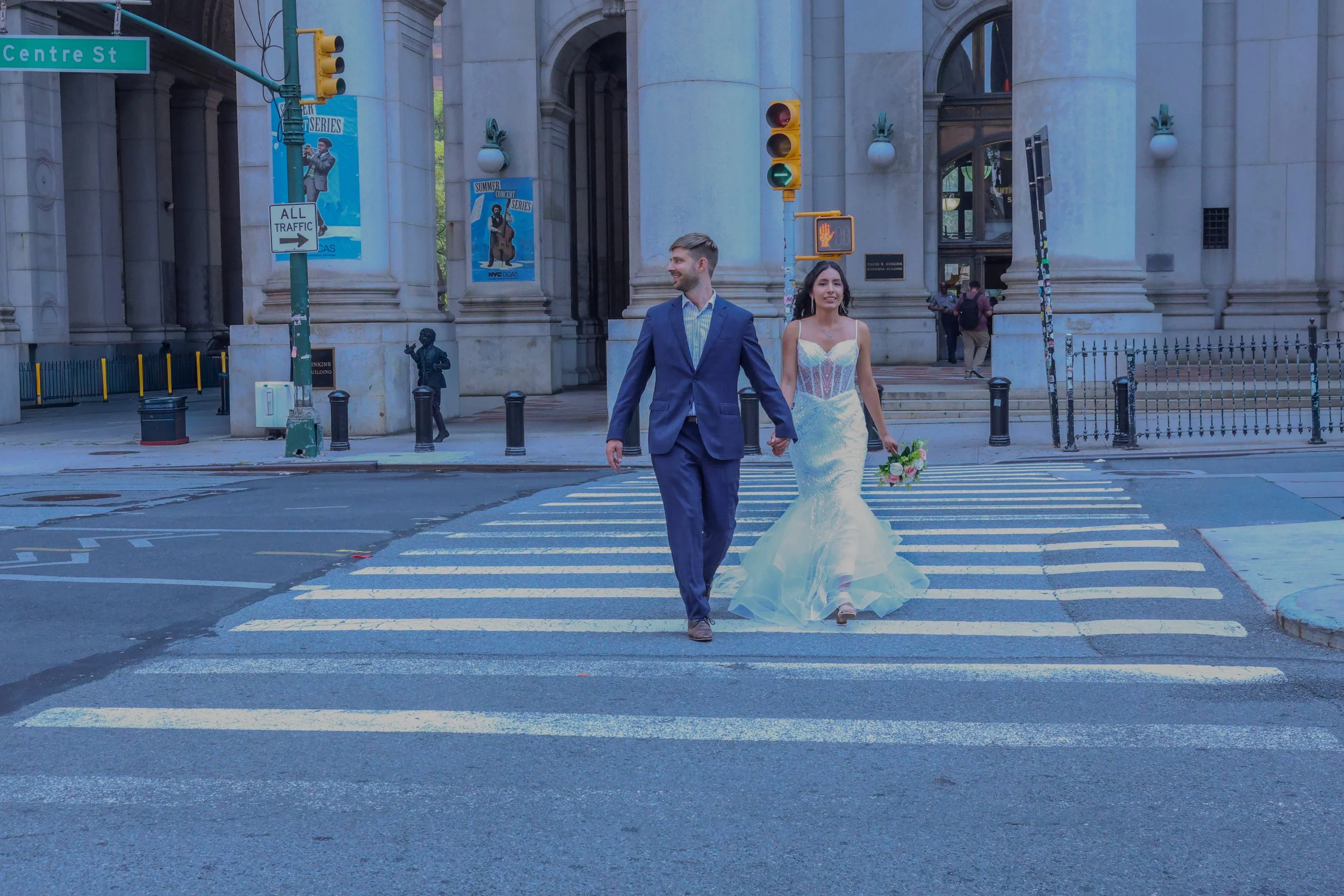 A newlywed couple holding hands and walking across a crosswalk in front of a large historic building. The groom is wearing a blue suit and the bride is in a white wedding dress holding a bouquet of flowers. Traffic lights and city signs are visible i