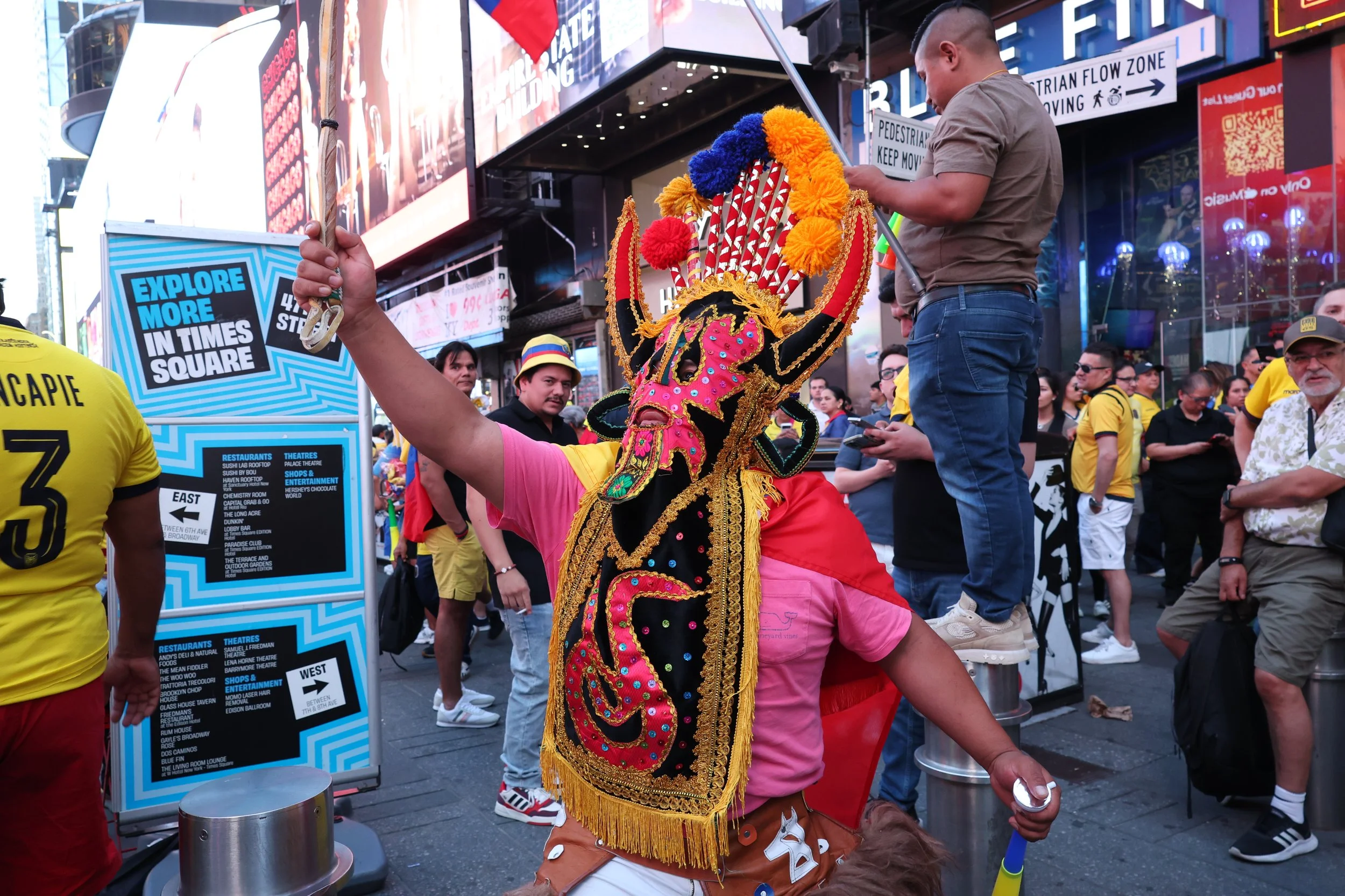 A performer dressed in a colorful, ornate costume with a large, elaborate headpiece performing in Times Square surrounded by a crowd of people and various signs and billboards.