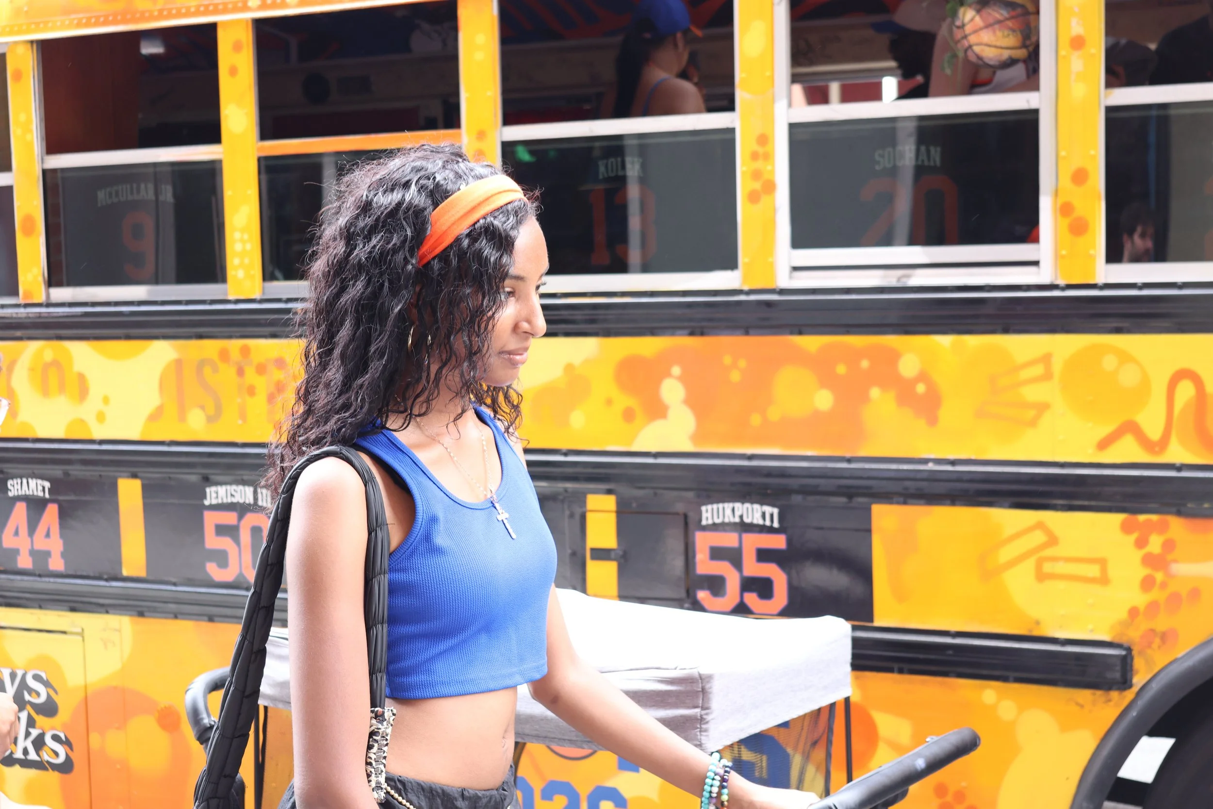 A woman with curly dark hair, wearing an orange headband and a blue crop top, is standing near a yellow food truck decorated with orange patterns. She is holding the handle of a wheelchair and has multiple bracelets on her left wrist.