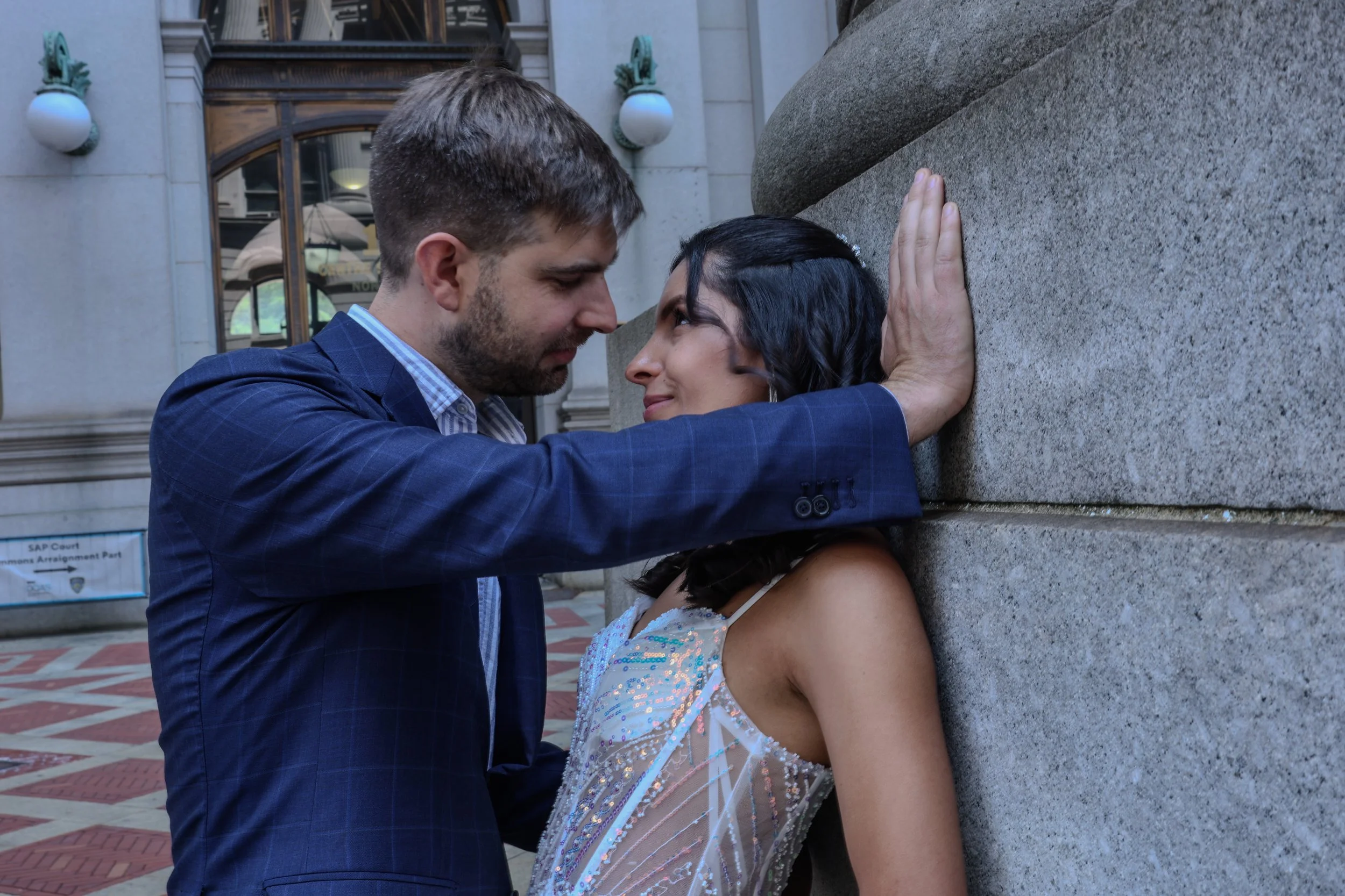 A man in a blue suit and a woman in a decorative dress lean against a large stone wall, face to face with foreheads touching, in an outdoor urban setting.
