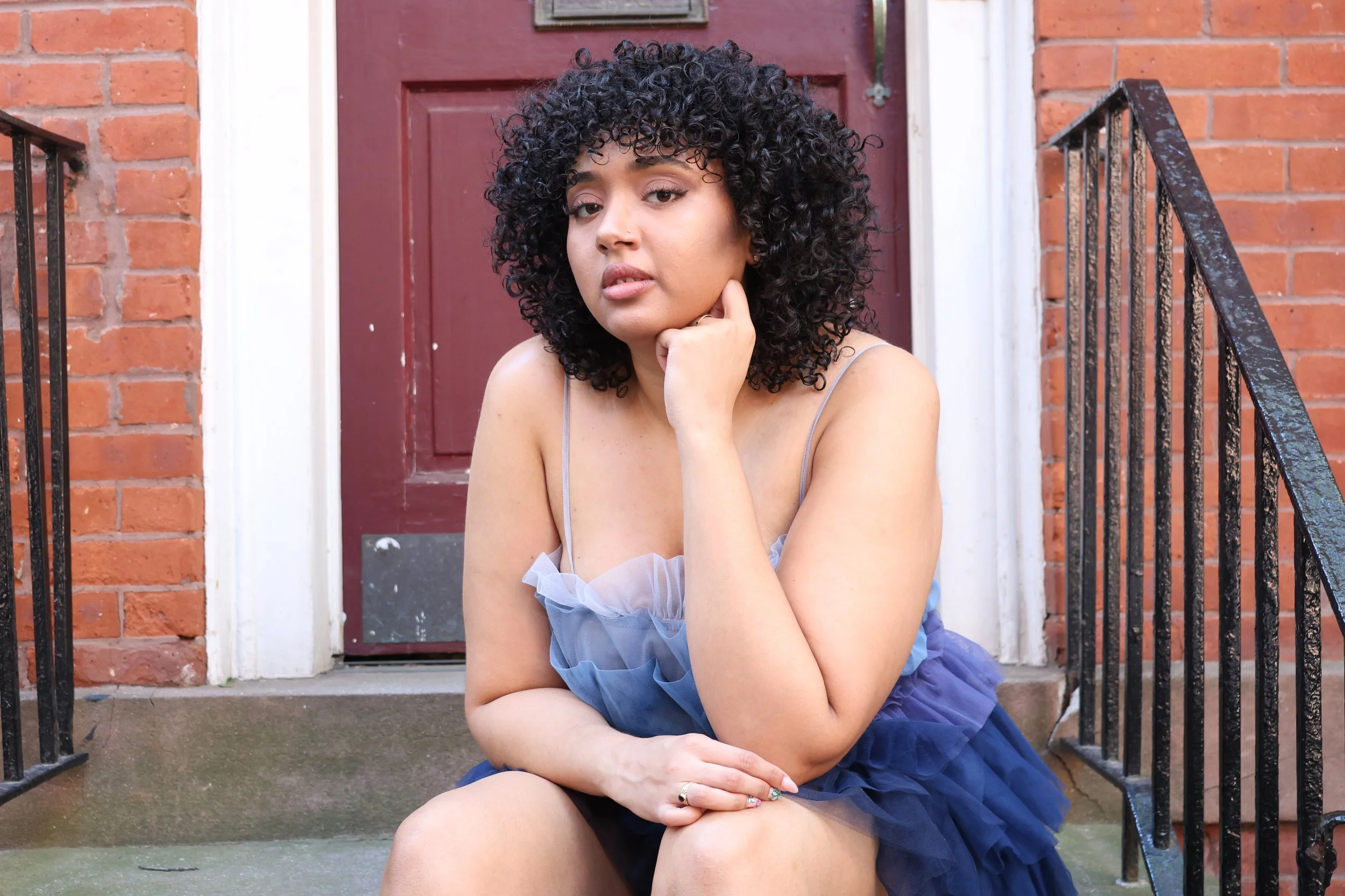 A woman with curly black hair sitting on concrete stairs in front of a brick building with a red door. She is wearing a blue dress with thin straps and has her left elbow resting on her knee while her hand touches her chin.