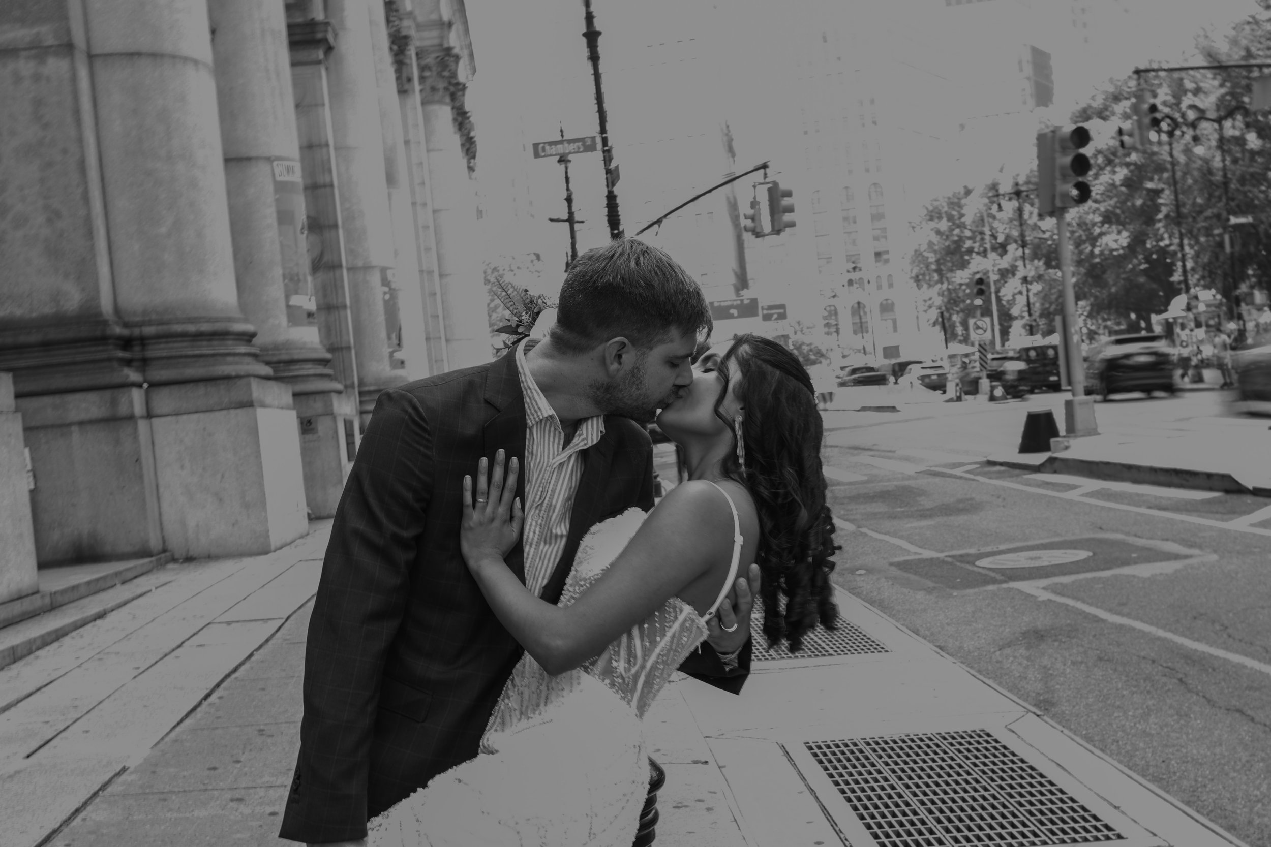 A black and white photo of a loving couple kissing on a city street with traffic lights and tall buildings in the background.