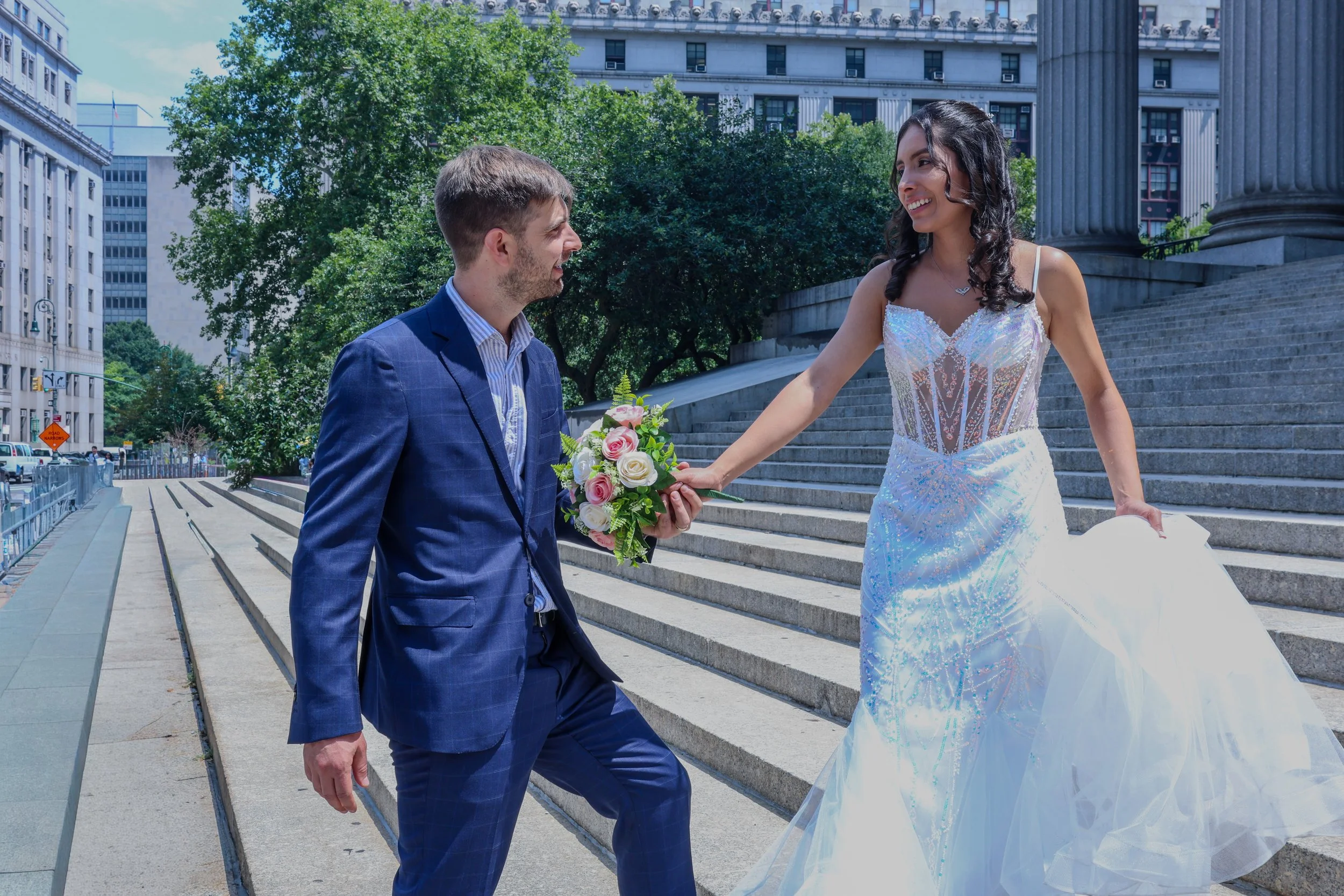 A couple dressed in wedding attire exchanging a bouquet of pink and white roses outdoors on steps in a city with tall buildings and trees.