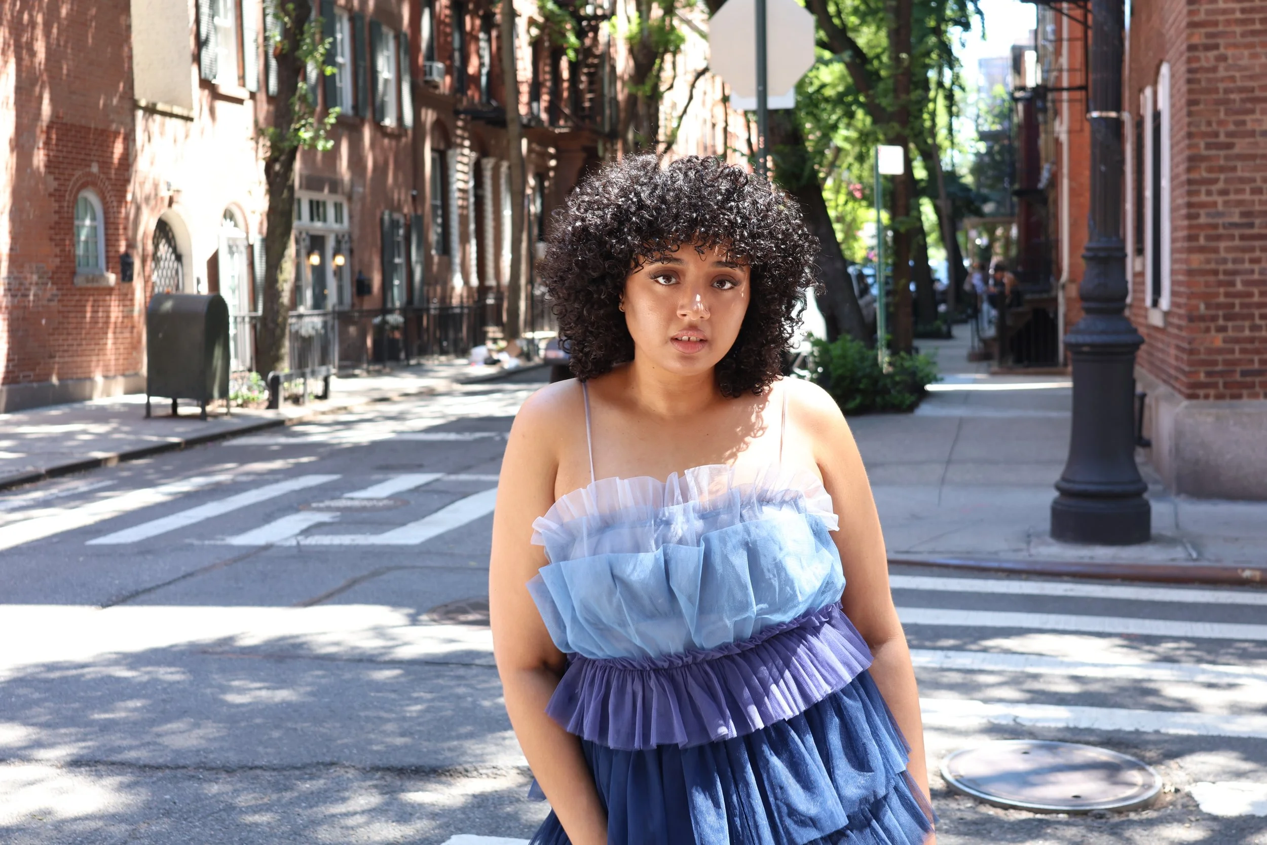 Young woman with curly hair wearing a blue dress standing on a city sidewalk in front of buildings and trees, with sunlight casting shadows.