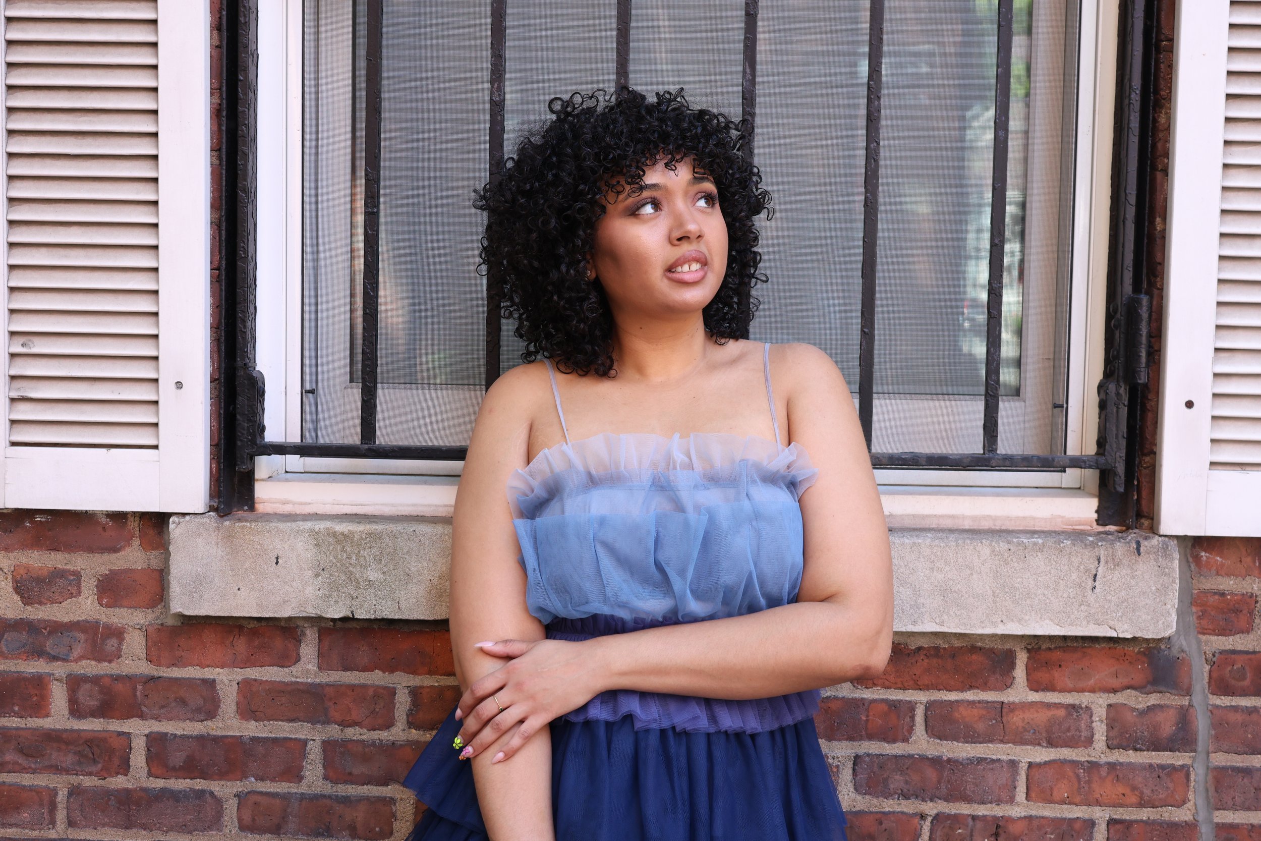 A young woman with curly black hair and light brown skin standing in front of a brick wall and a window with shutters, wearing a blue dress, looking up with crossed arms.