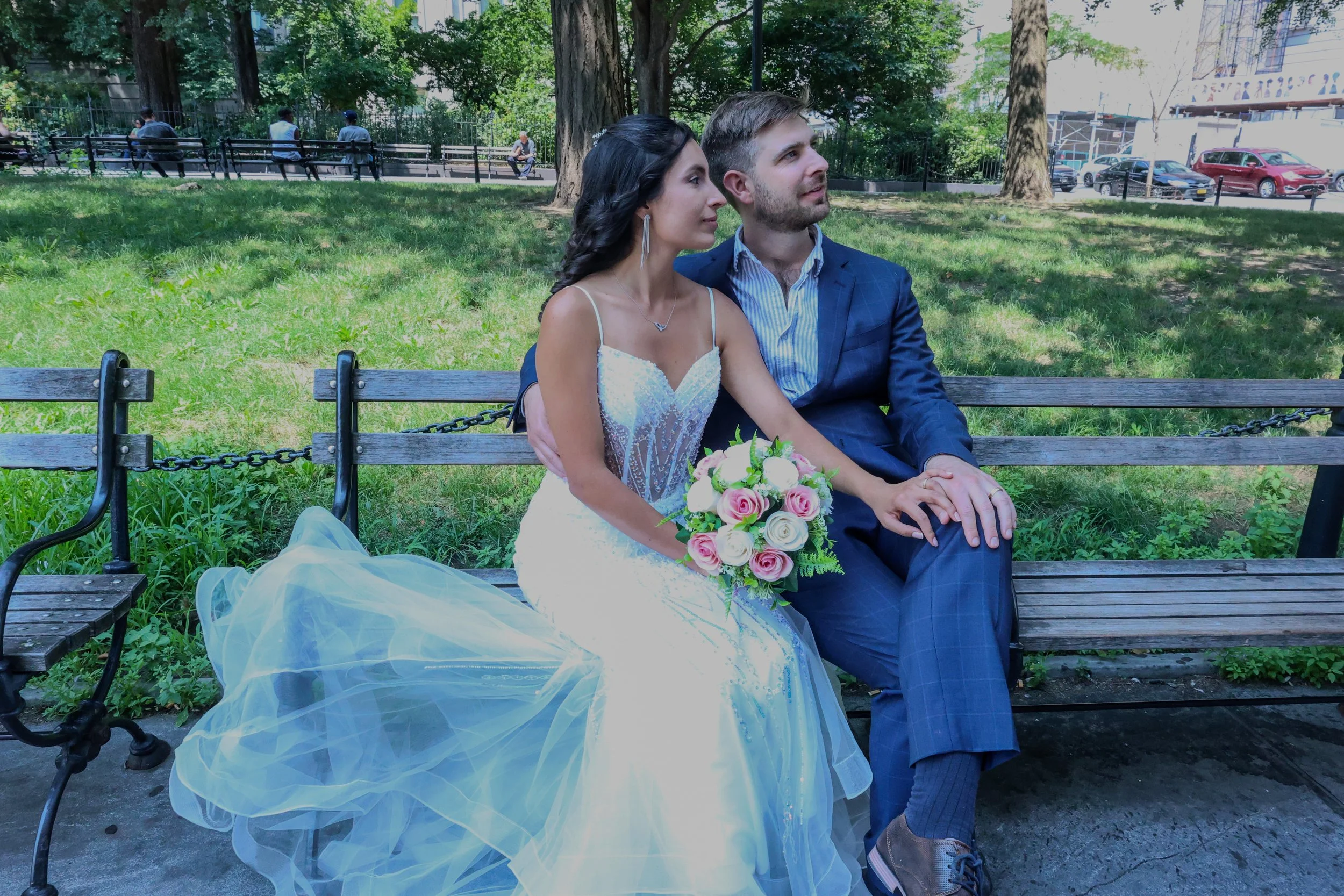 A bride and groom sitting on a park bench during daytime, with the bride holding a bouquet of pink and white roses, both looking content, amid green trees and urban surroundings.