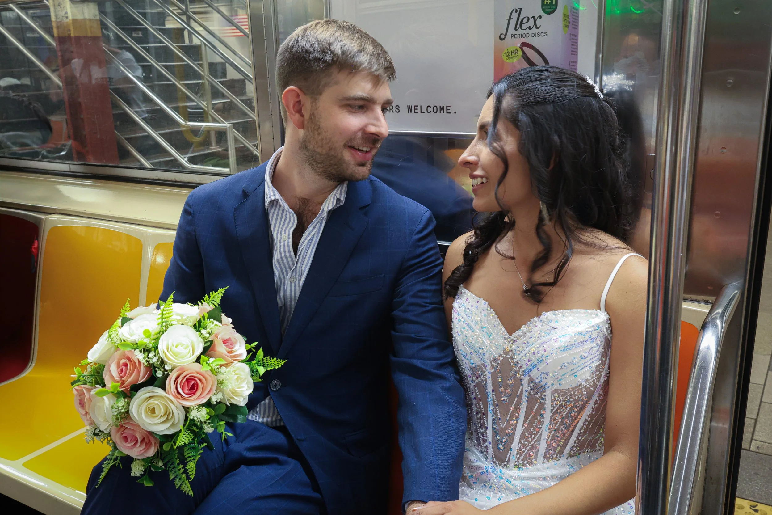 A newlywed couple sitting close together on a subway, smiling and gazing into each other's eyes. The groom is holding a bouquet of pink and white roses with green foliage. They are dressed in formal wedding attire, with the bride wearing a sparkling 