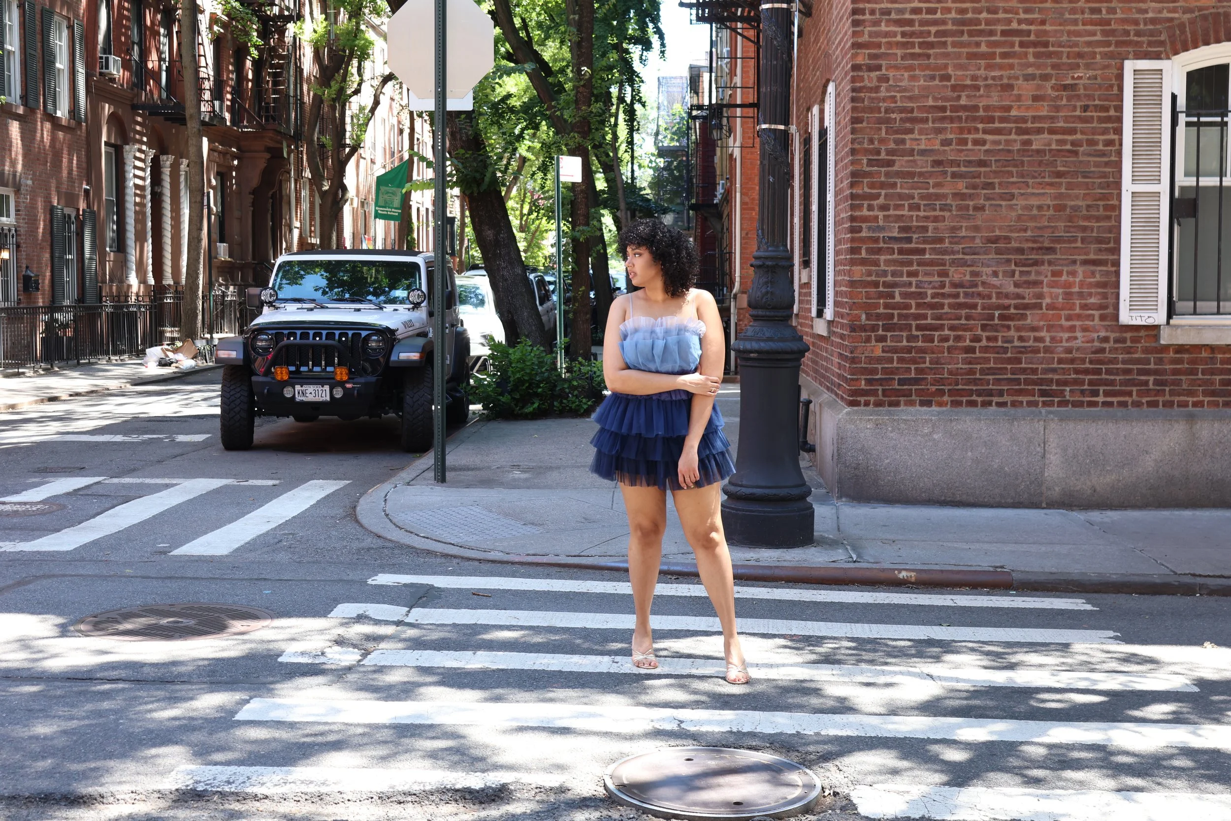 A woman in a blue layered dress and nude high heels crossing a city street at a crosswalk. She has curly black hair and is standing near a black lamppost, with a row of red brick buildings and parked cars in the background. The street is shaded by tr