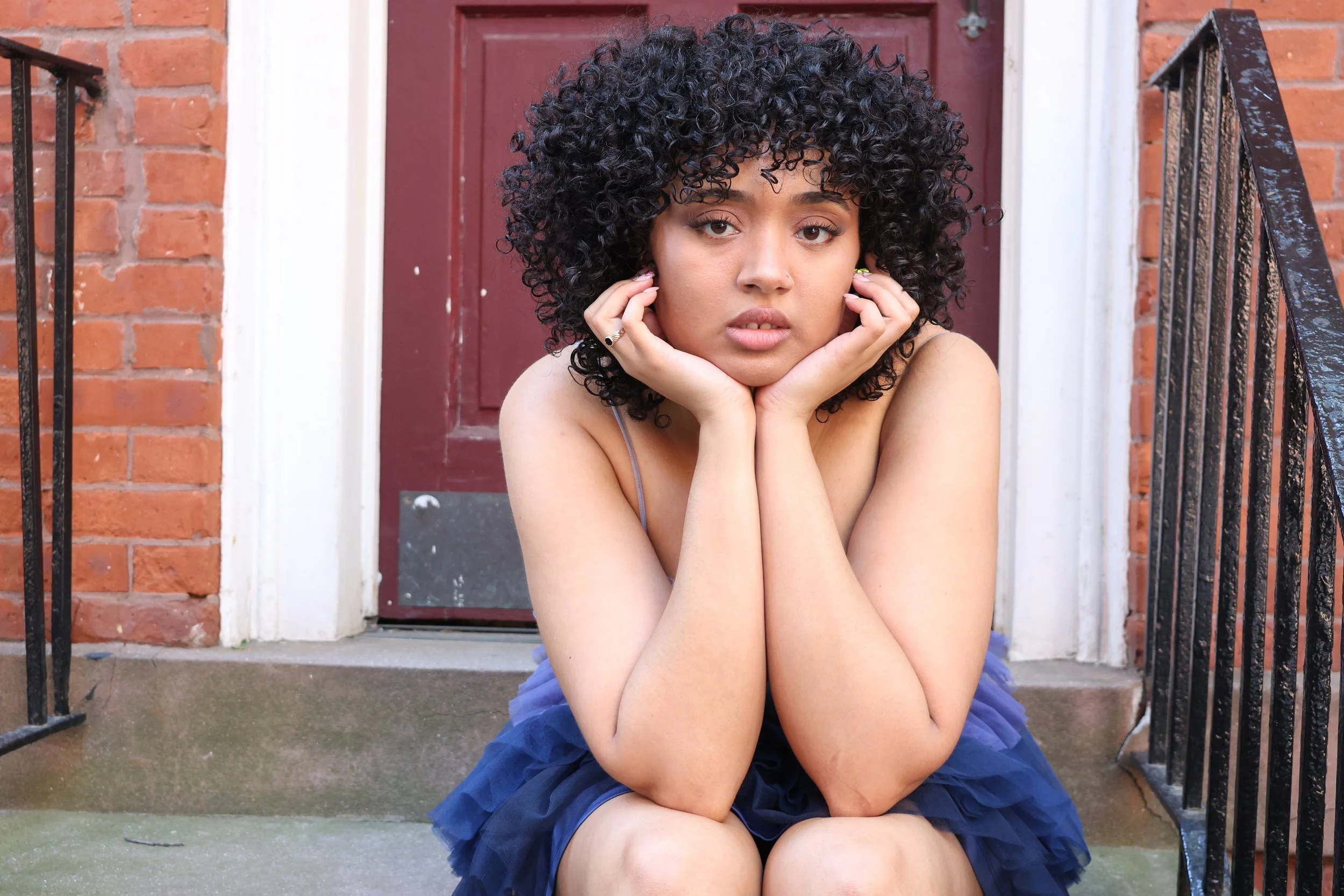 Young woman with curly black hair sitting on doorstep in front of a brick house with a red door, looking at the camera with her hands resting on her face.