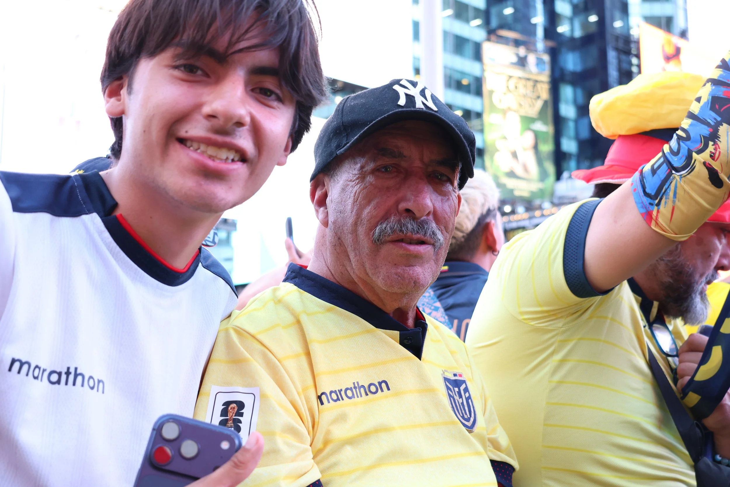 Two men in yellow sports shirts and a woman in a white sports shirt smiling and taking selfies during a public event in an urban setting.