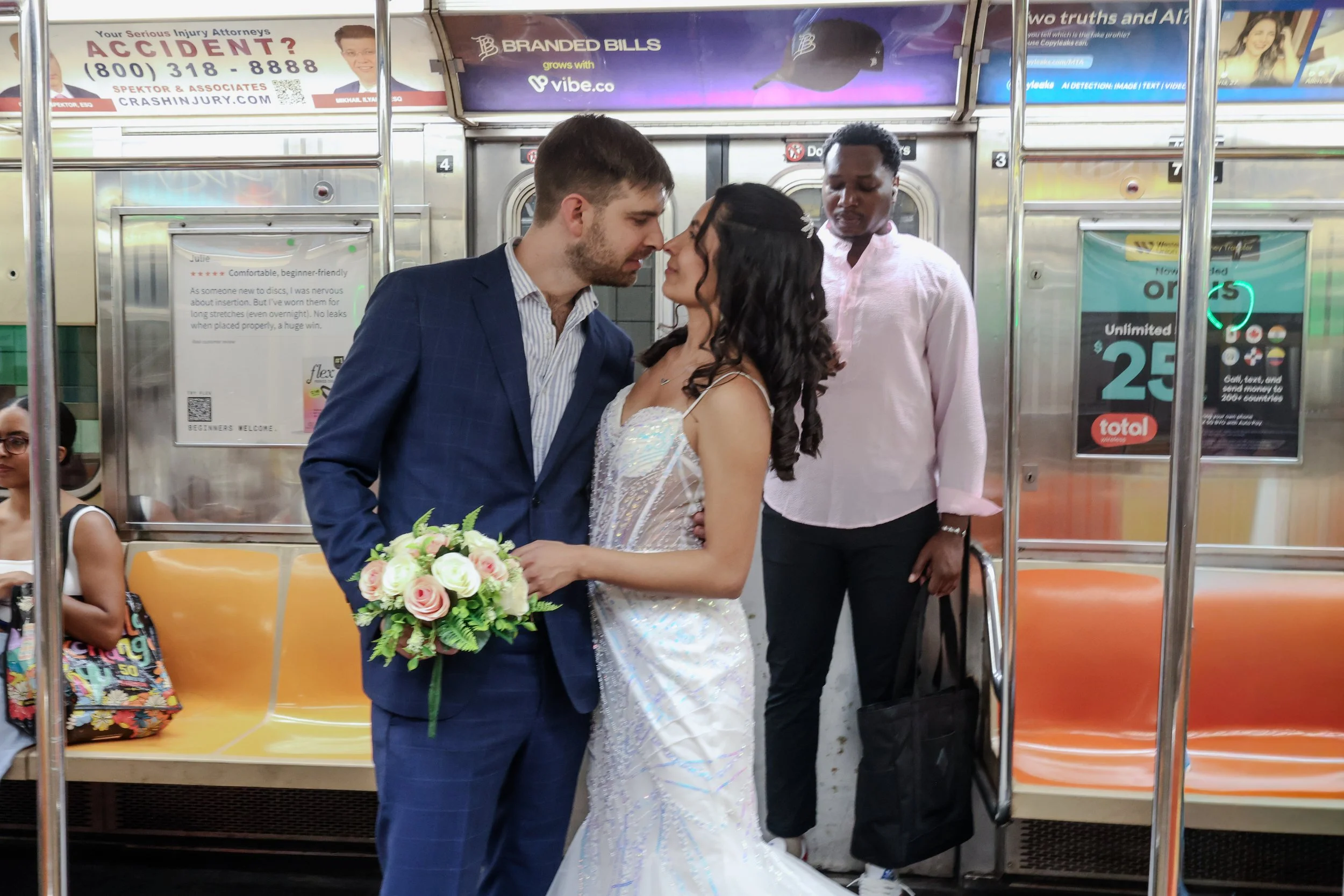A couple dressed in wedding attire standing close together in a subway train station, about to kiss. The woman holds a bouquet of flowers. There are two other women sitting on the train in the background, one with glasses and a colorful dress, and th