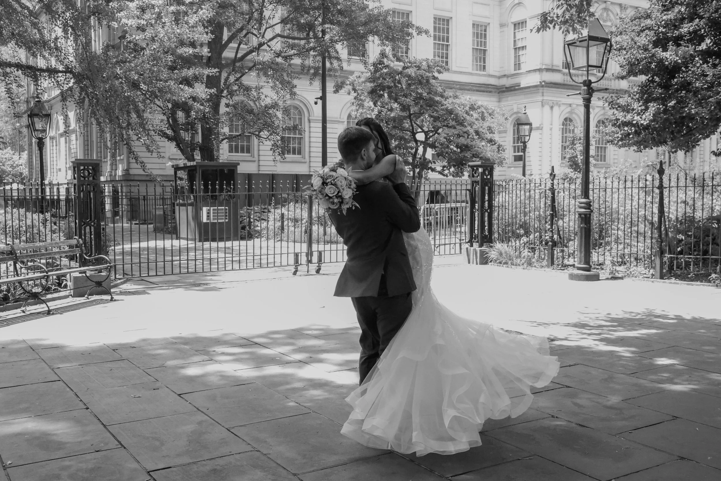 A couple dressed in wedding attire, embracing and dancing outdoors in front of a historic building with large windows and trees, during daytime.