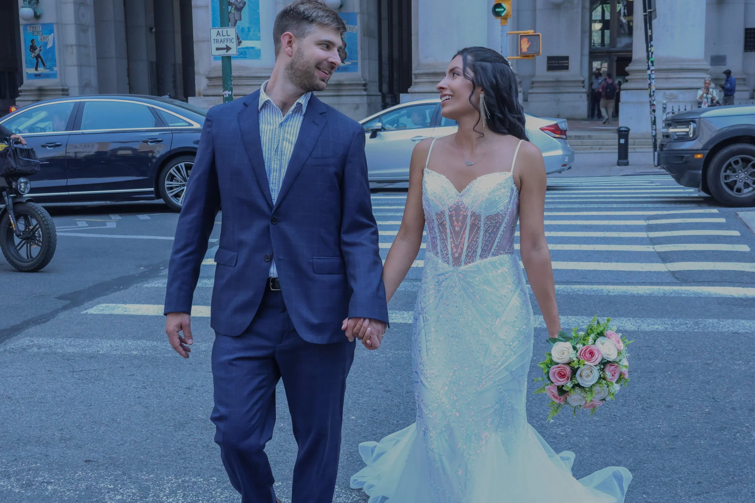 A newlywed couple holding hands and smiling at each other while walking across a city street, with cars and pedestrians in the background.