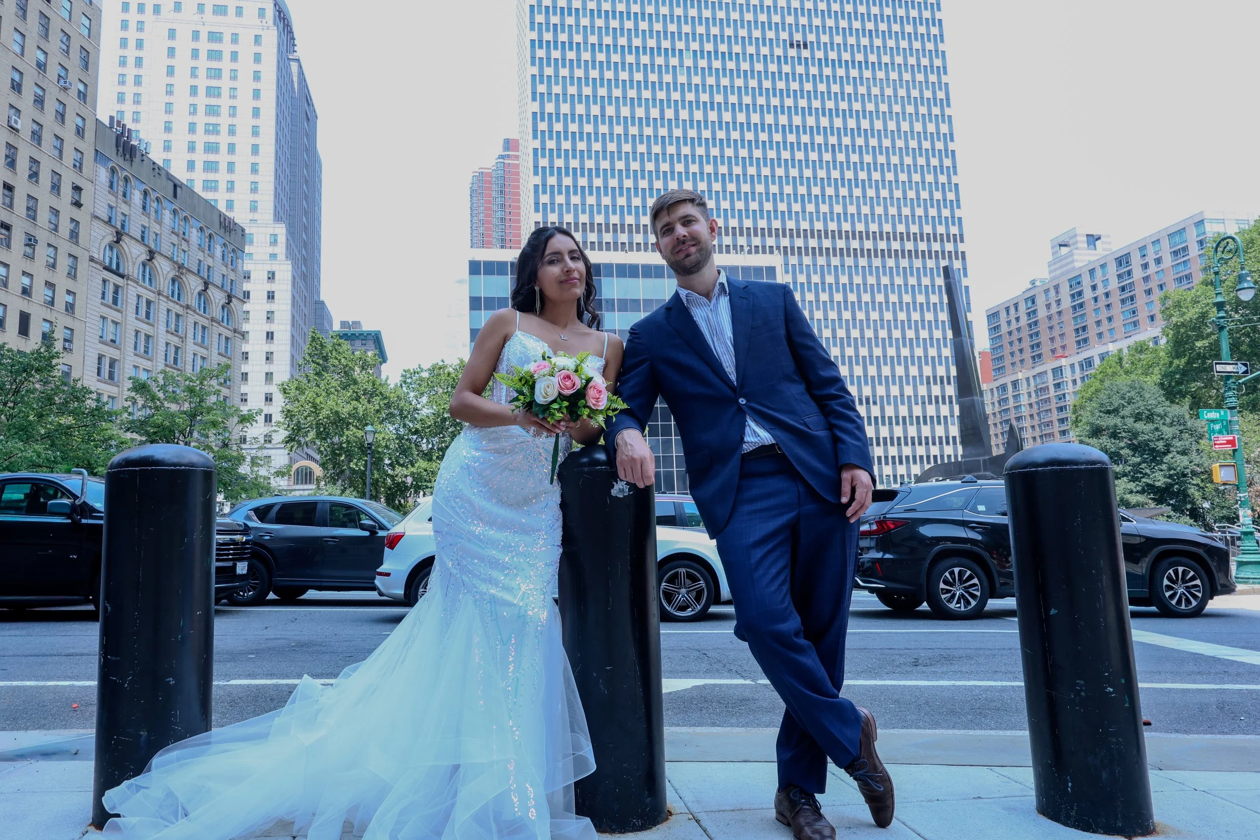 A couple dressed in wedding attire posing in an urban setting with tall buildings and parked cars, standing behind a black bollard.