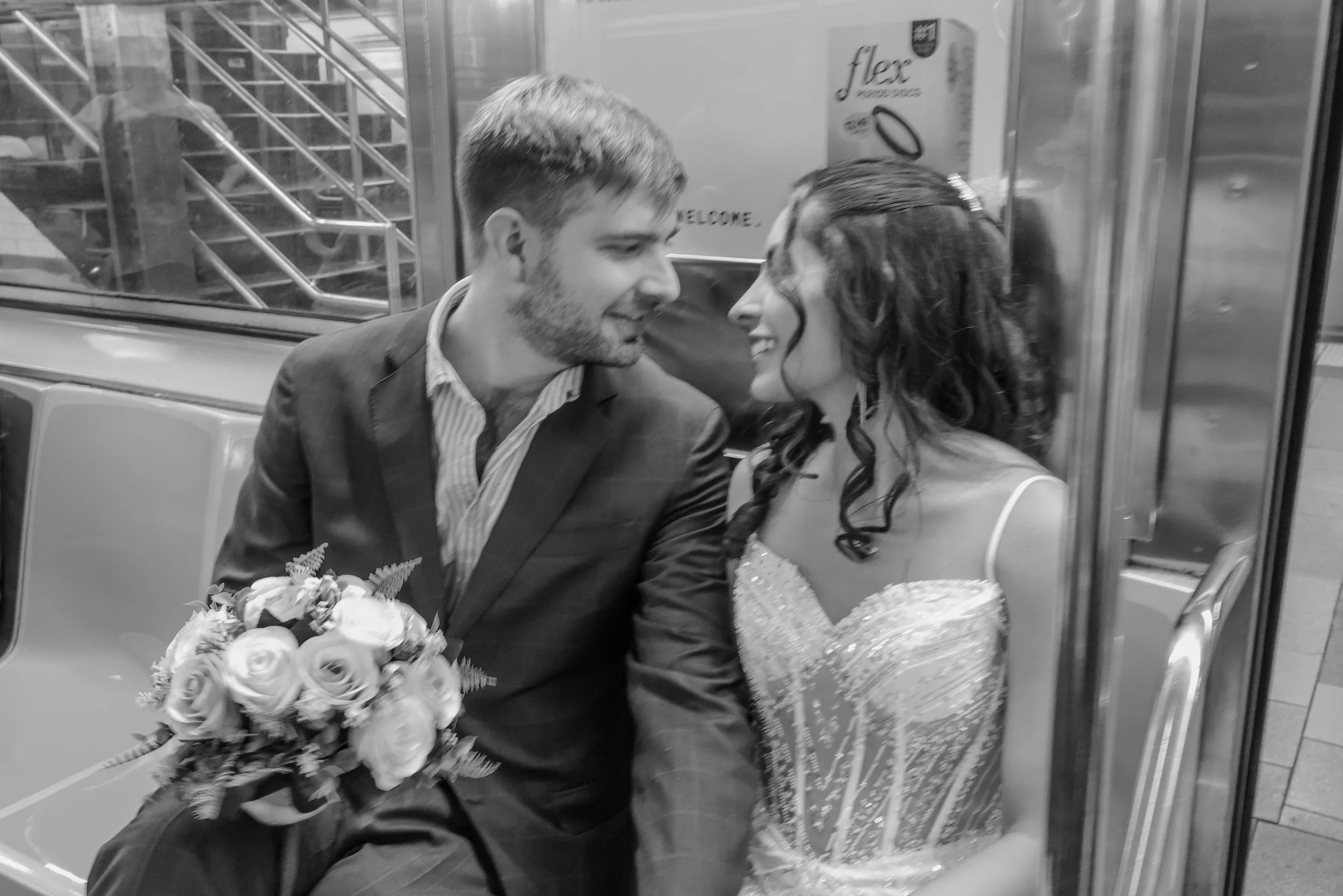 A man and woman with smiles are sitting close together on a subway train, with the woman holding a bouquet of flowers and the man holding a bouquet. The woman is dressed for a formal occasion, possibly a wedding, with dark curls and a dress with embe