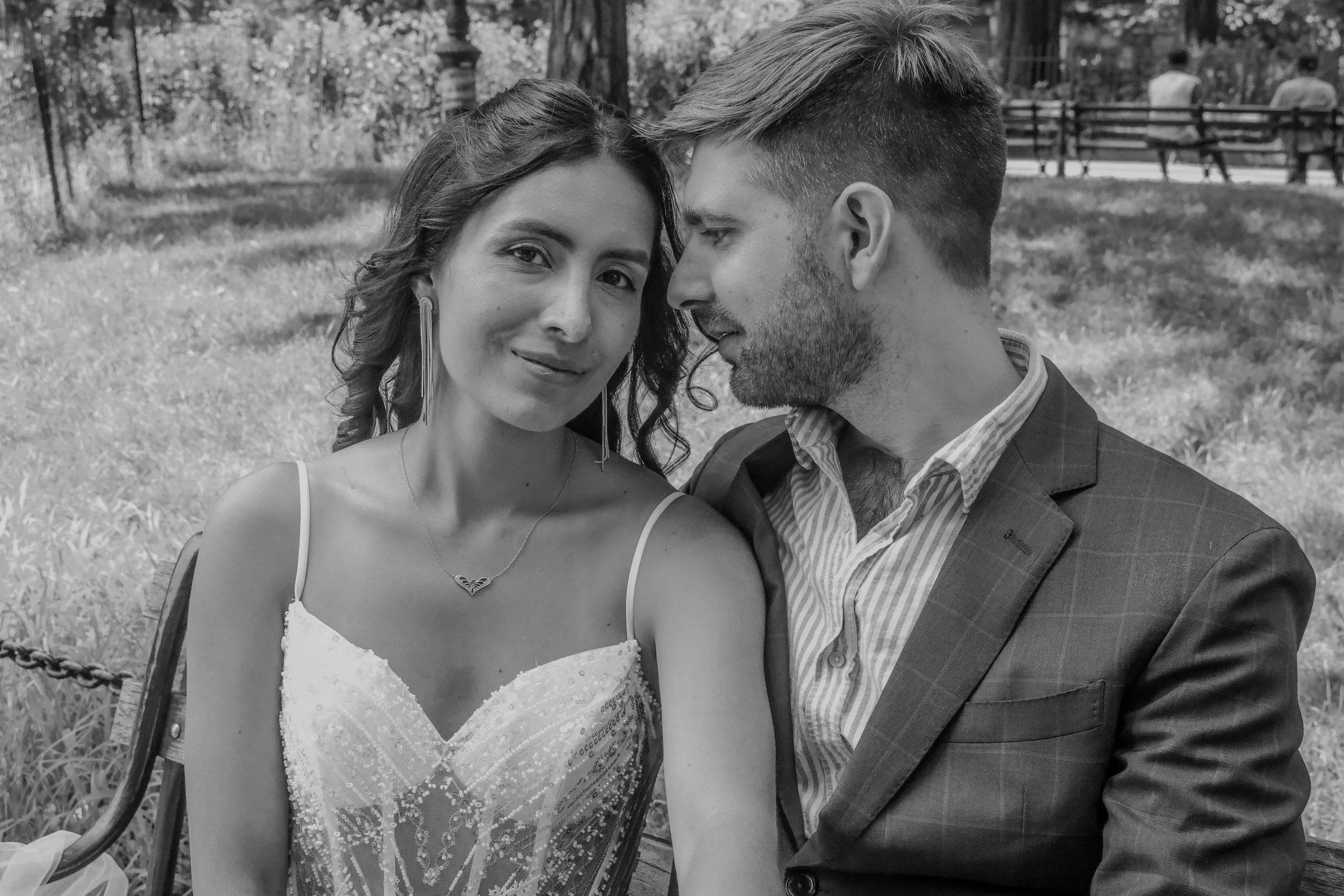 A black and white photo of a couple in a park, close together, with the woman looking at the camera and the man facing her with him eyes closed, sitting on a wooden bench with trees and a fence in the background.