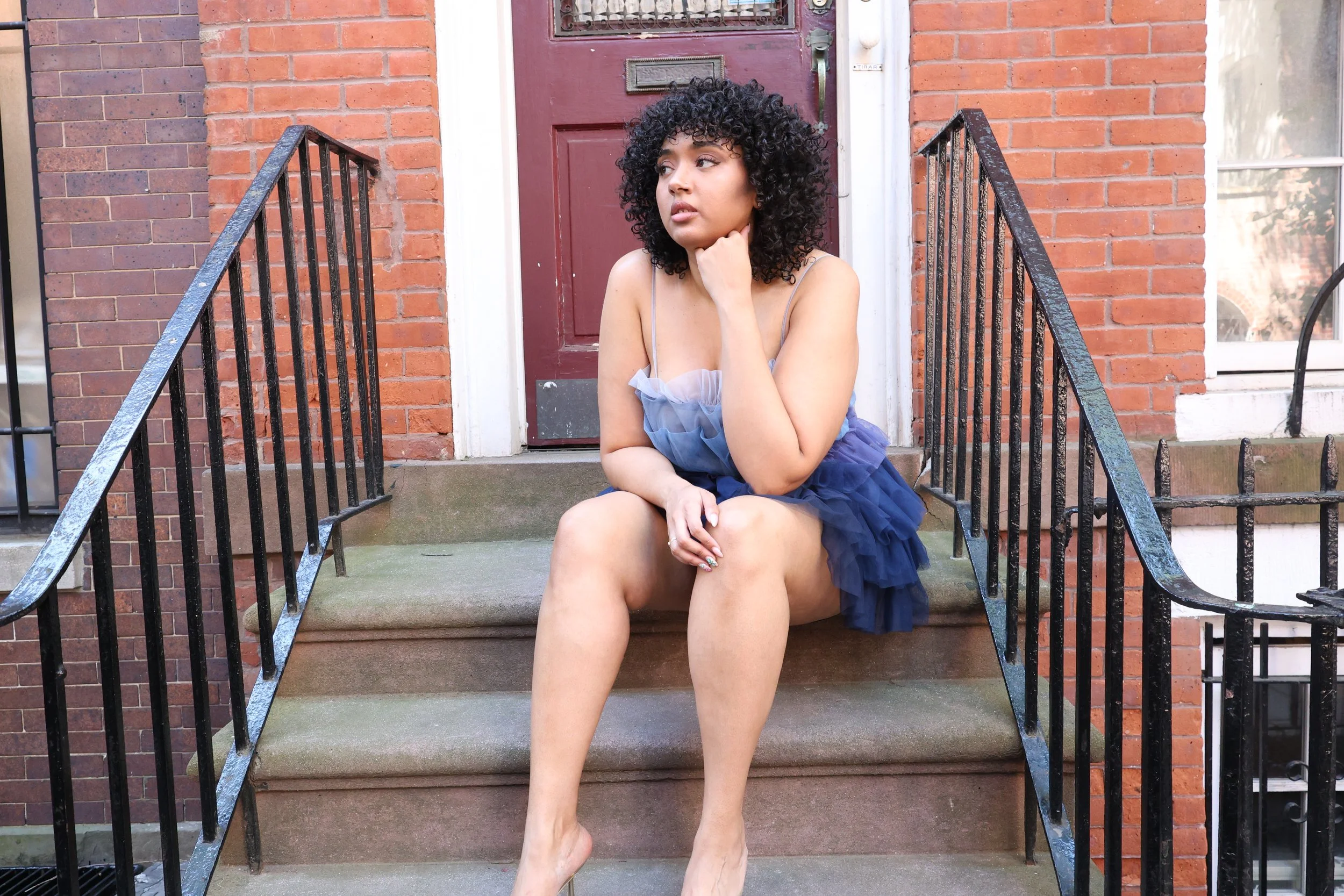 A woman with curly black hair sitting on the steps outside a brick building, wearing a gray tank top and a navy blue skirt, looking to the side with her right hand resting on her neck.