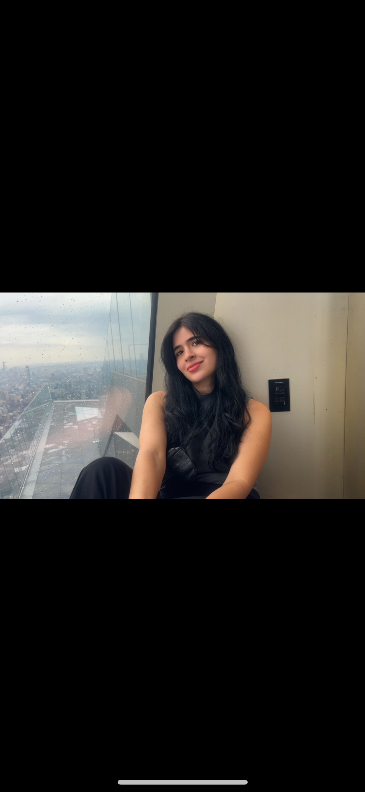 A woman with long black hair and a black sleeveless top sitting indoors near a large window on a rainy day, with a city skyline in the background.