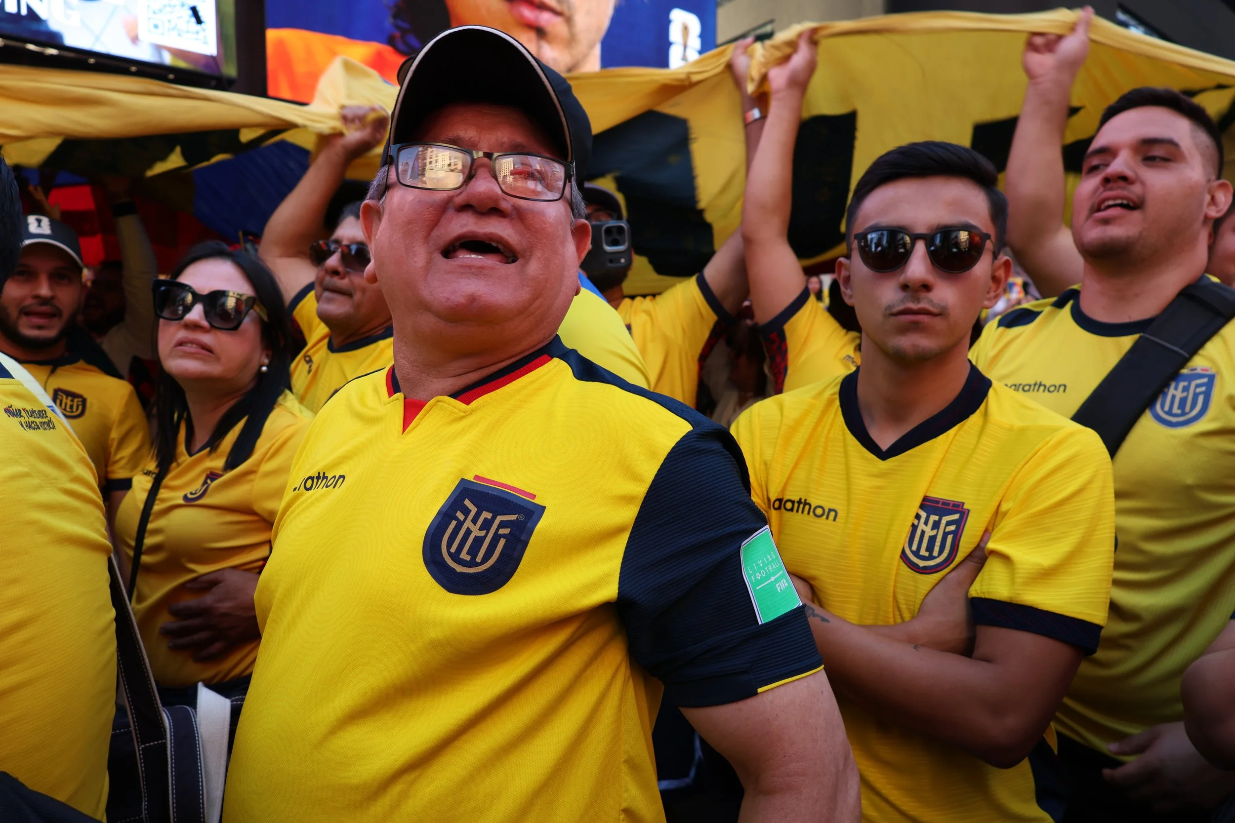 Group of soccer fans wearing yellow jerseys, some with sunglasses, gathered outdoors. One man in the front is speaking or singing, while others are standing close together, some holding a yellow and black flag or scarf above their heads.