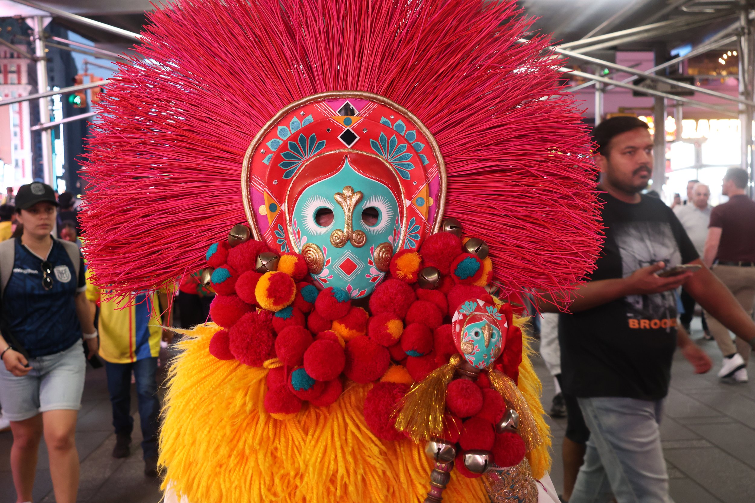 Person wearing a colorful traditional costume with a decorated mask, surrounded by bright red and yellow feathers, during a cultural event or festival.