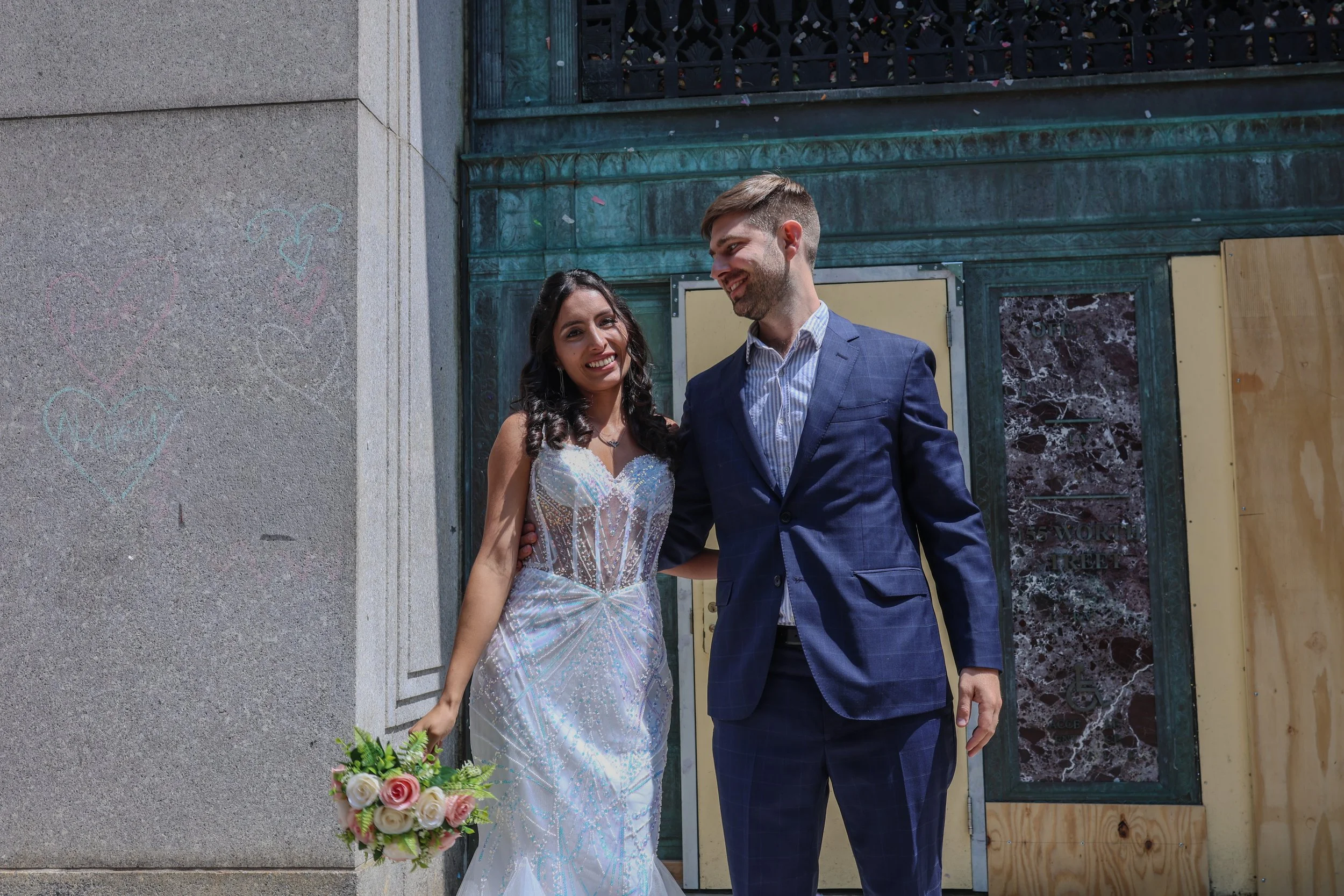 A newlywed couple, a woman in a white wedding dress holding a bouquet of flowers and a man in a blue suit, standing outside near a building with graffiti hearts on the wall, smiling and looking at each other.