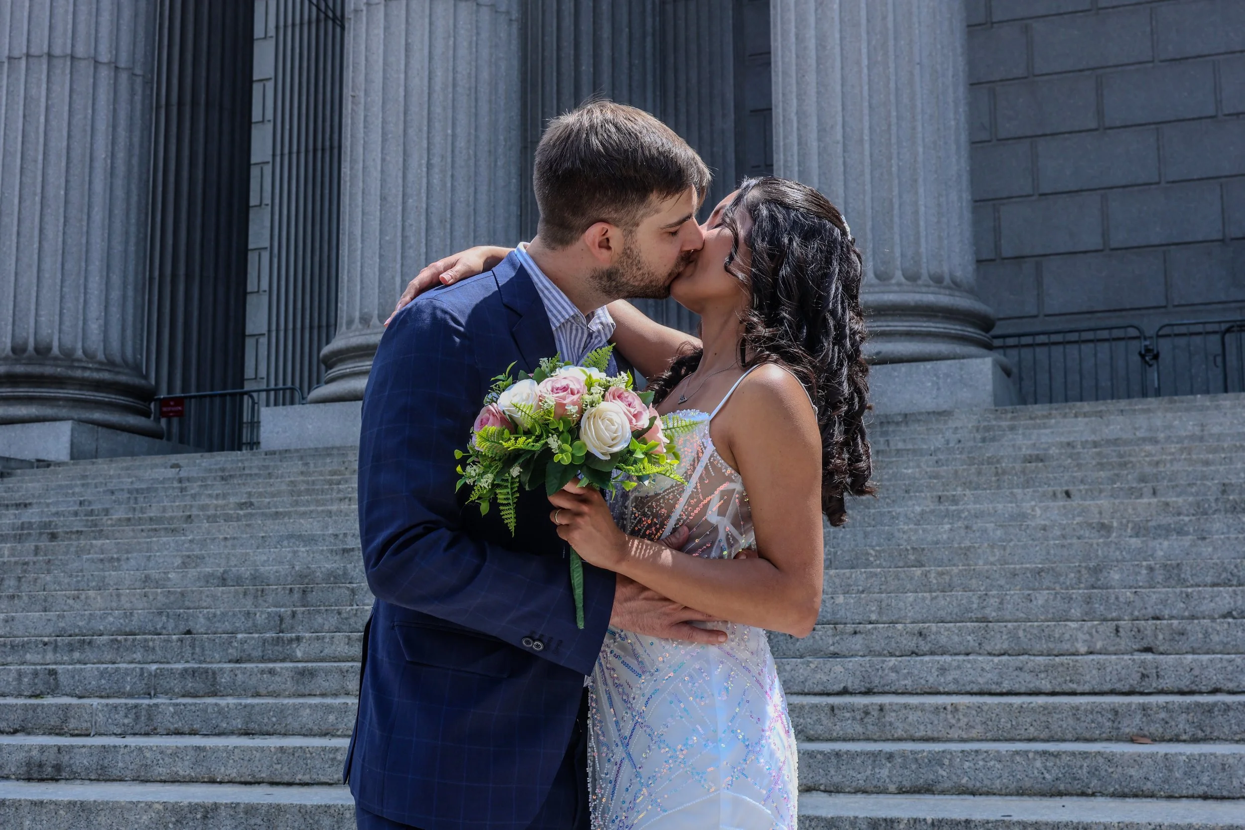 A couple sharing a kiss on the steps of a government building, with the woman holding a bouquet of pink and white flowers.