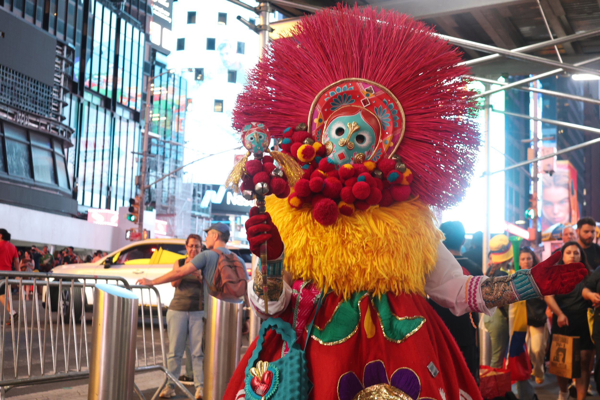 Person dressed in a colorful traditional costume with a large, fluffy red headdress, decorated mask, and vibrant clothing, participating in a street parade or celebration in an urban area with buildings, traffic, and crowd in the background.
