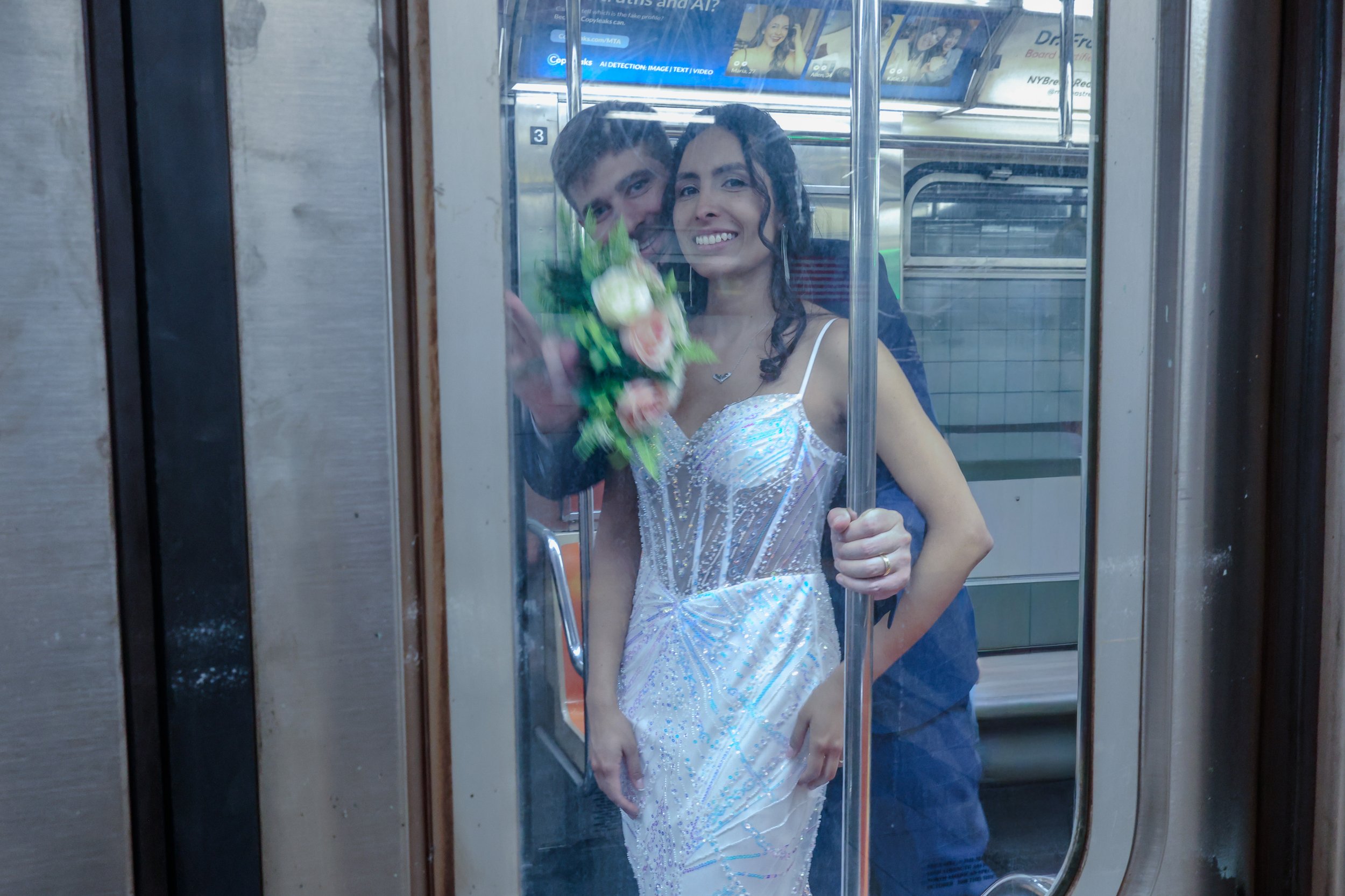 A bride in a sparkly white wedding dress holding a bouquet, smiling at the camera, visible through the glass of an elevator door. A man stands behind her, slightly out of focus.