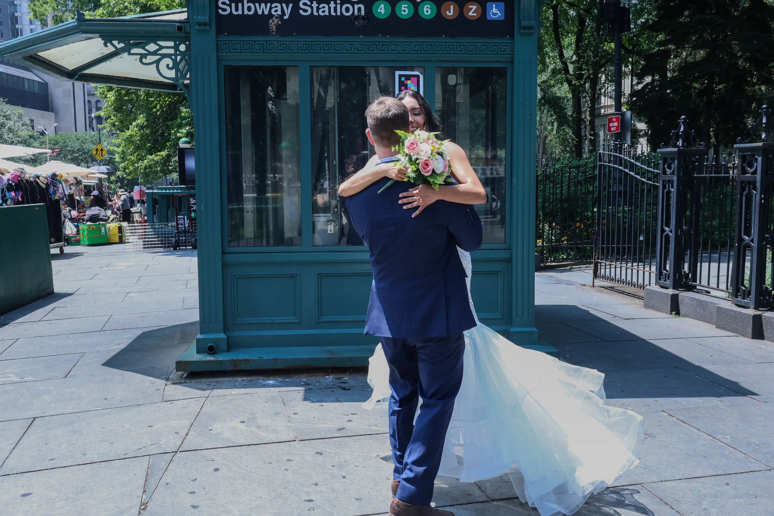 A couple embracing, with the woman holding a bouquet of flowers, standing outside a subway station entrance on a sunny day.