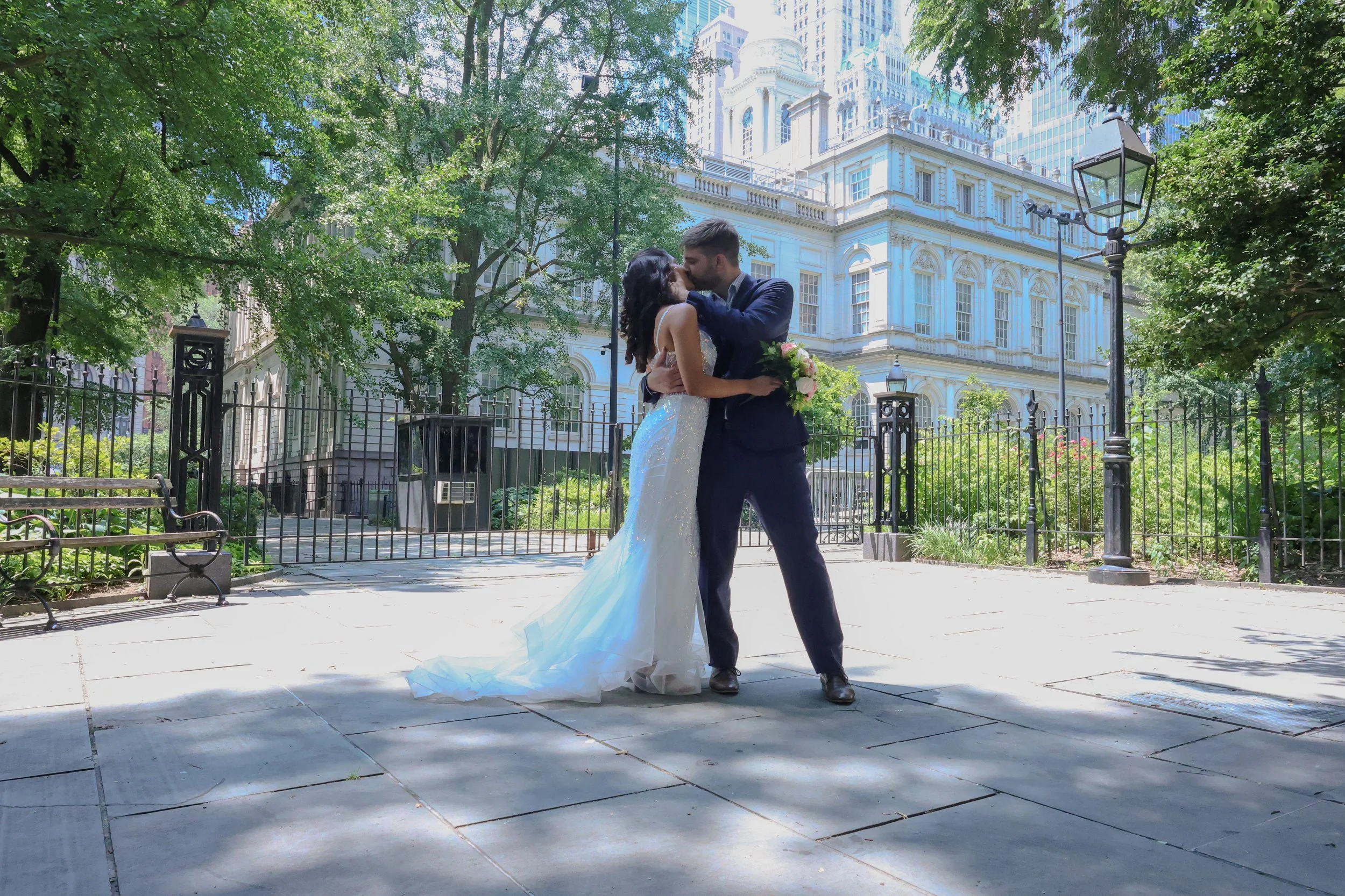 A newlywed couple sharing a kiss in a park with lush green trees, a white historic building in the background, and vintage lampposts.