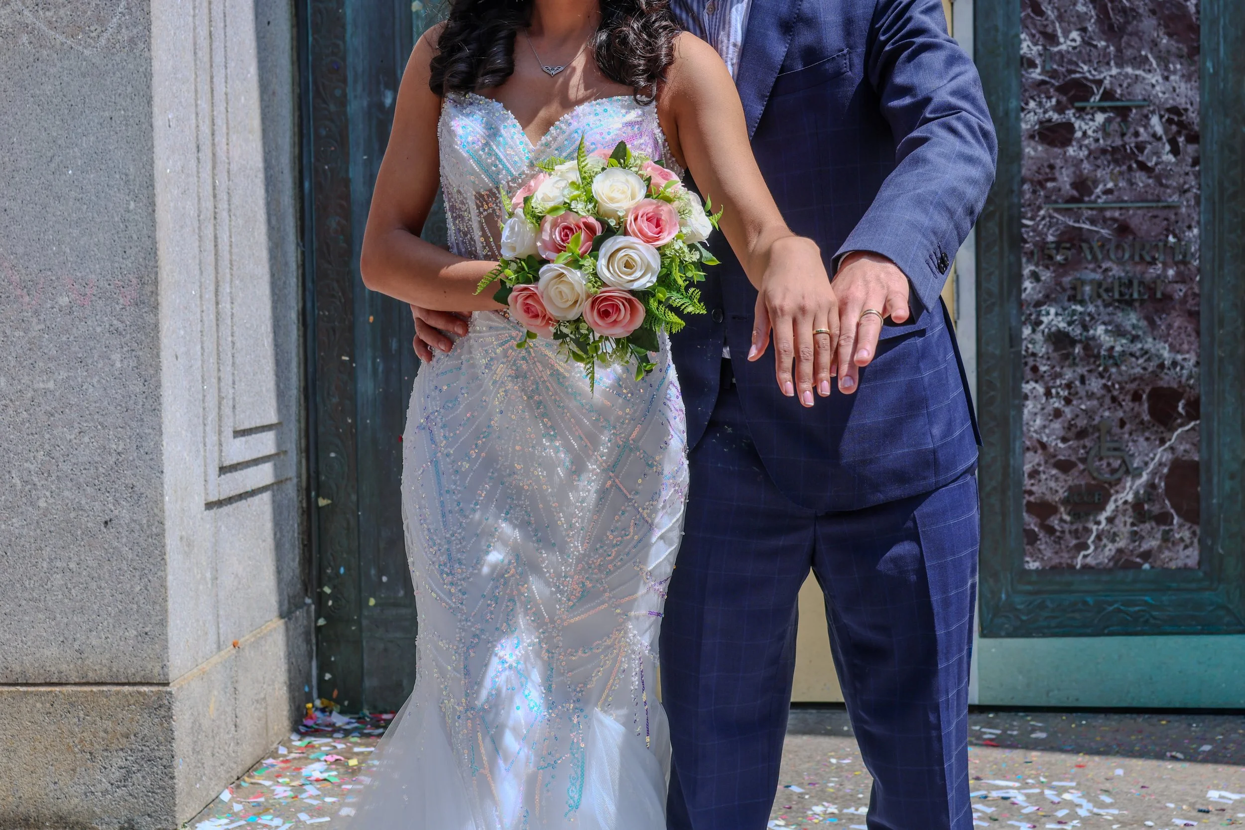 A bride and groom holding hands during their wedding ceremony outside. The bride is wearing a white, sparkly dress and holding a bouquet of pink and white roses. The groom is wearing a blue suit.