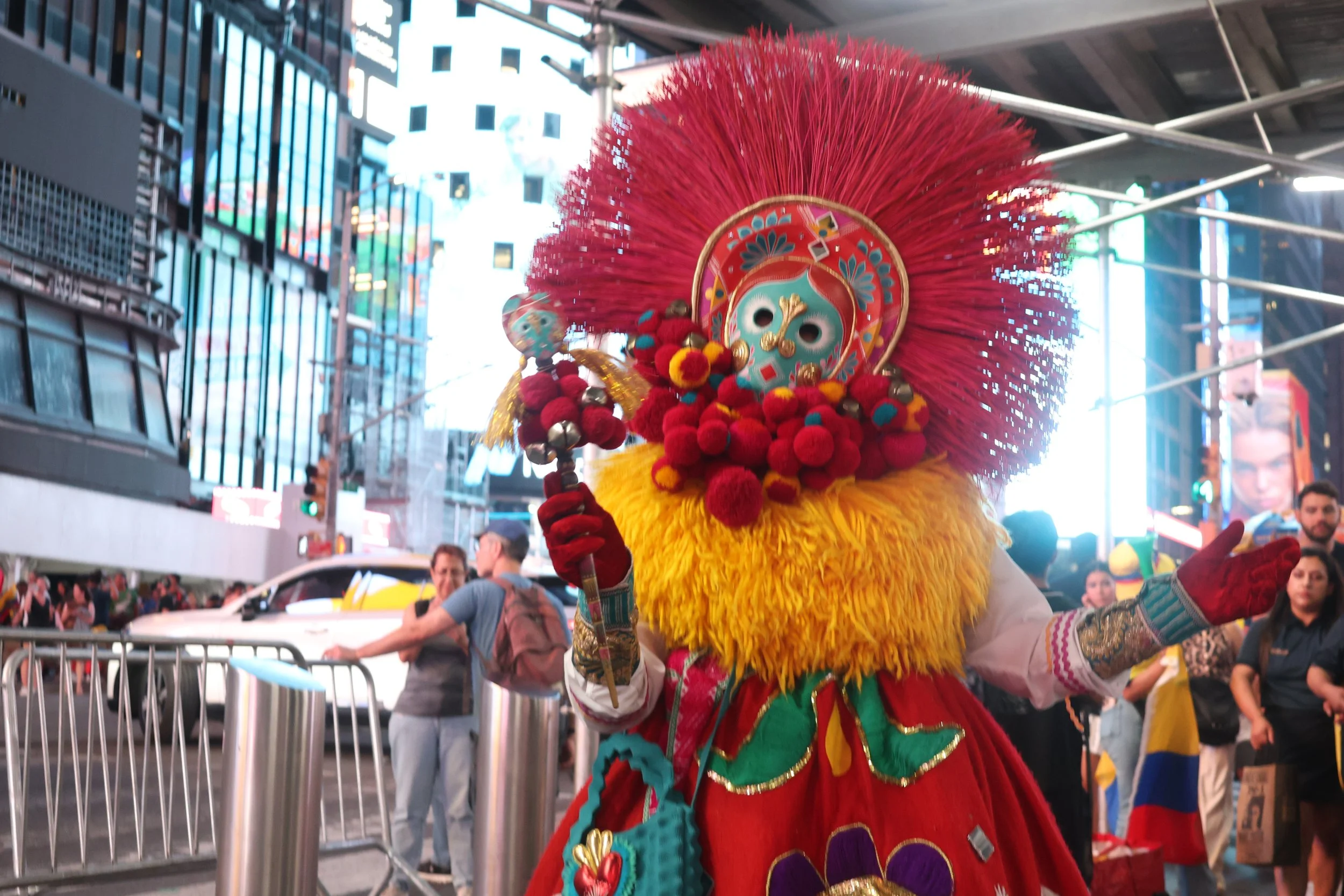 A performer dressed in a colorful costume with a large, red, feathered headdress performing in a busy city street with onlookers and tall buildings in the background.