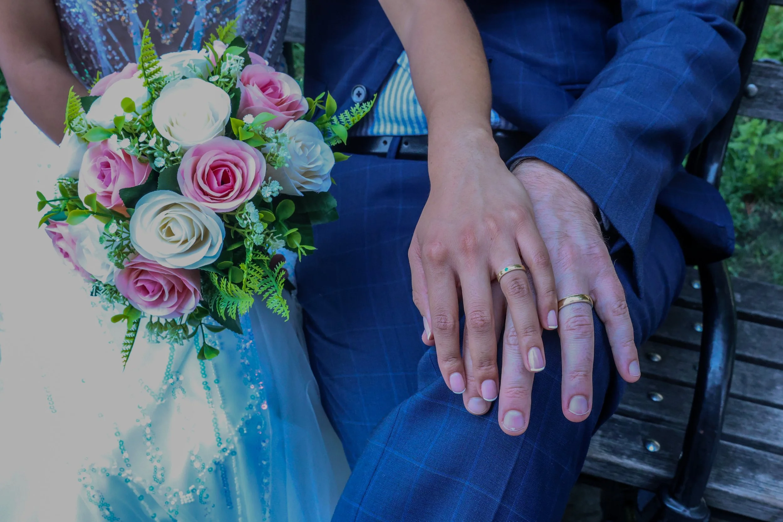 Close-up of a bride and groom sitting outdoors, with the bride holding a bouquet of pink and white roses and greenery, and the groom's hand resting on his knee, displaying wedding rings.