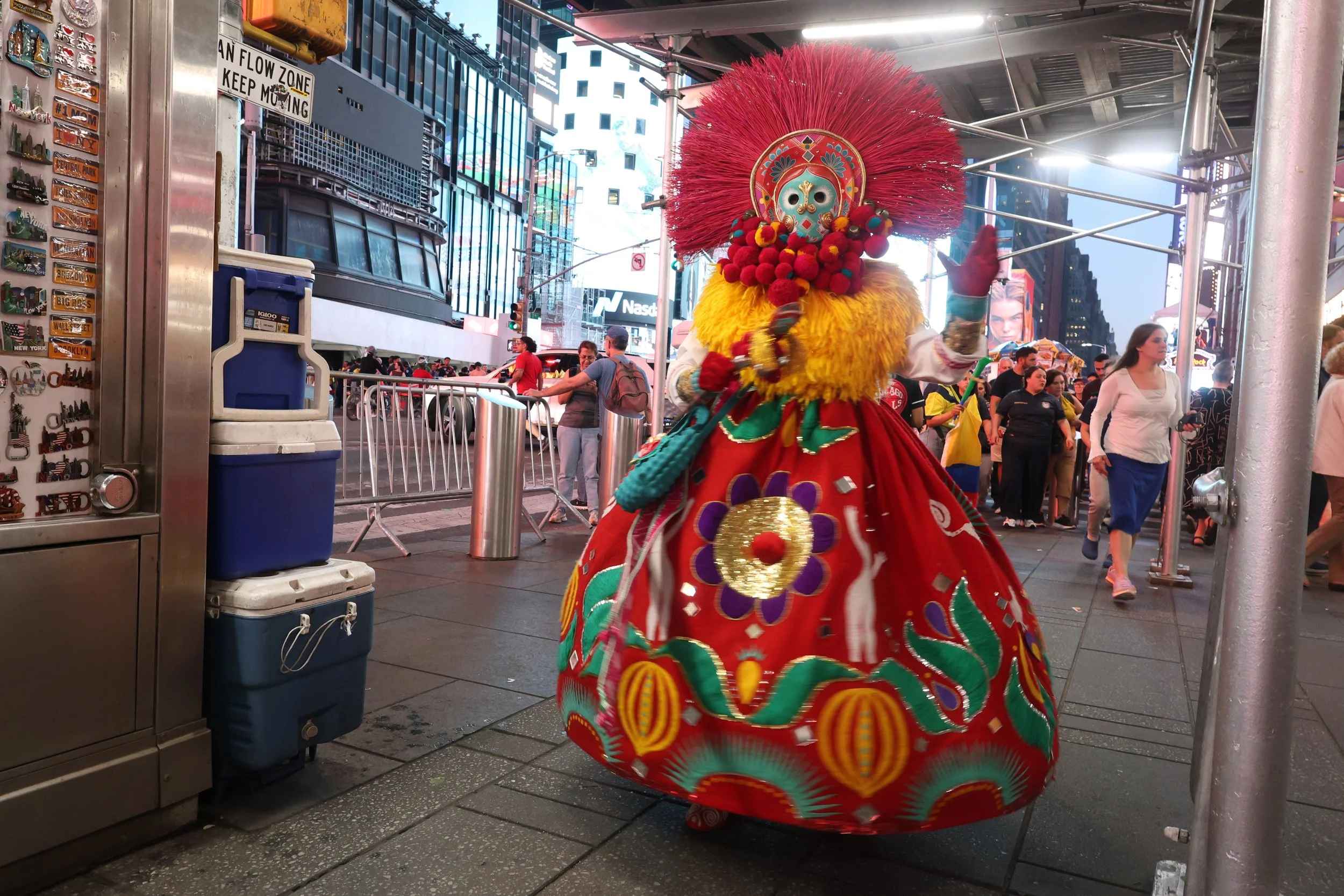 A performer in traditional Mexican attire, including a colorful embroidered dress and a large, ornate mask with red feathers, is seen in Times Square, New York City, surrounded by spectators and urban buildings.